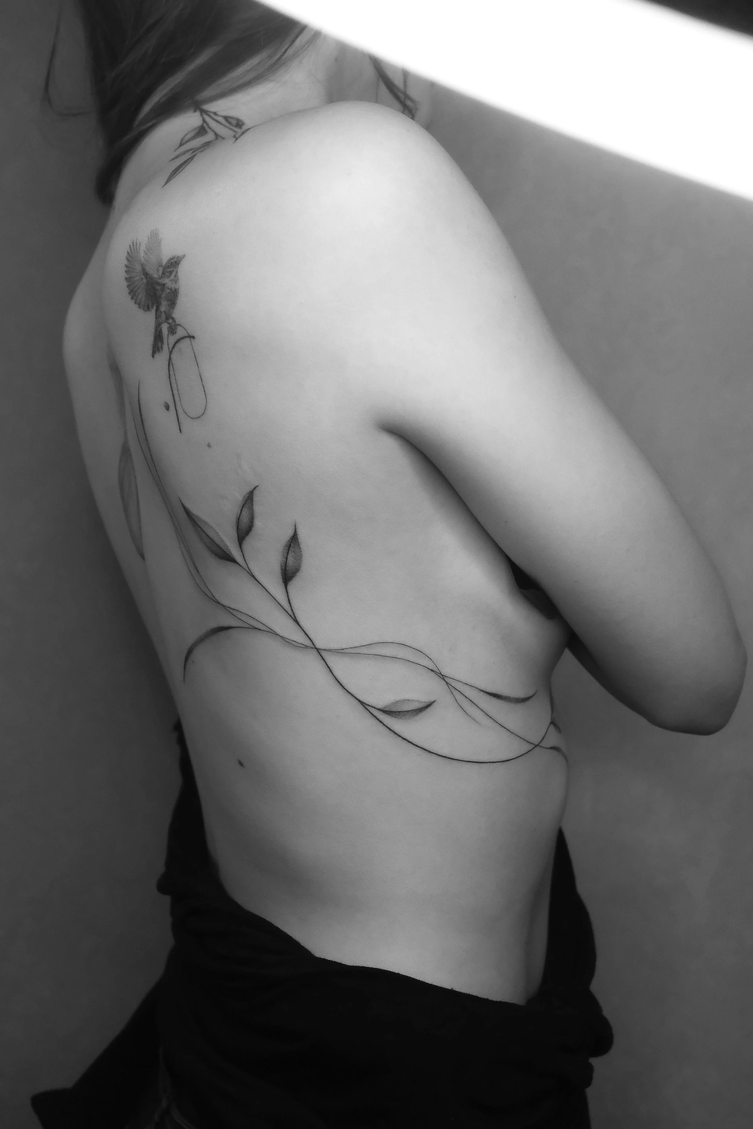 Black and white photo of a person with partial view of back and shoulder, showcasing a freehand tattoo done by Ola Getka in Aura Tattoo Reykjavik, wearing a black garment.