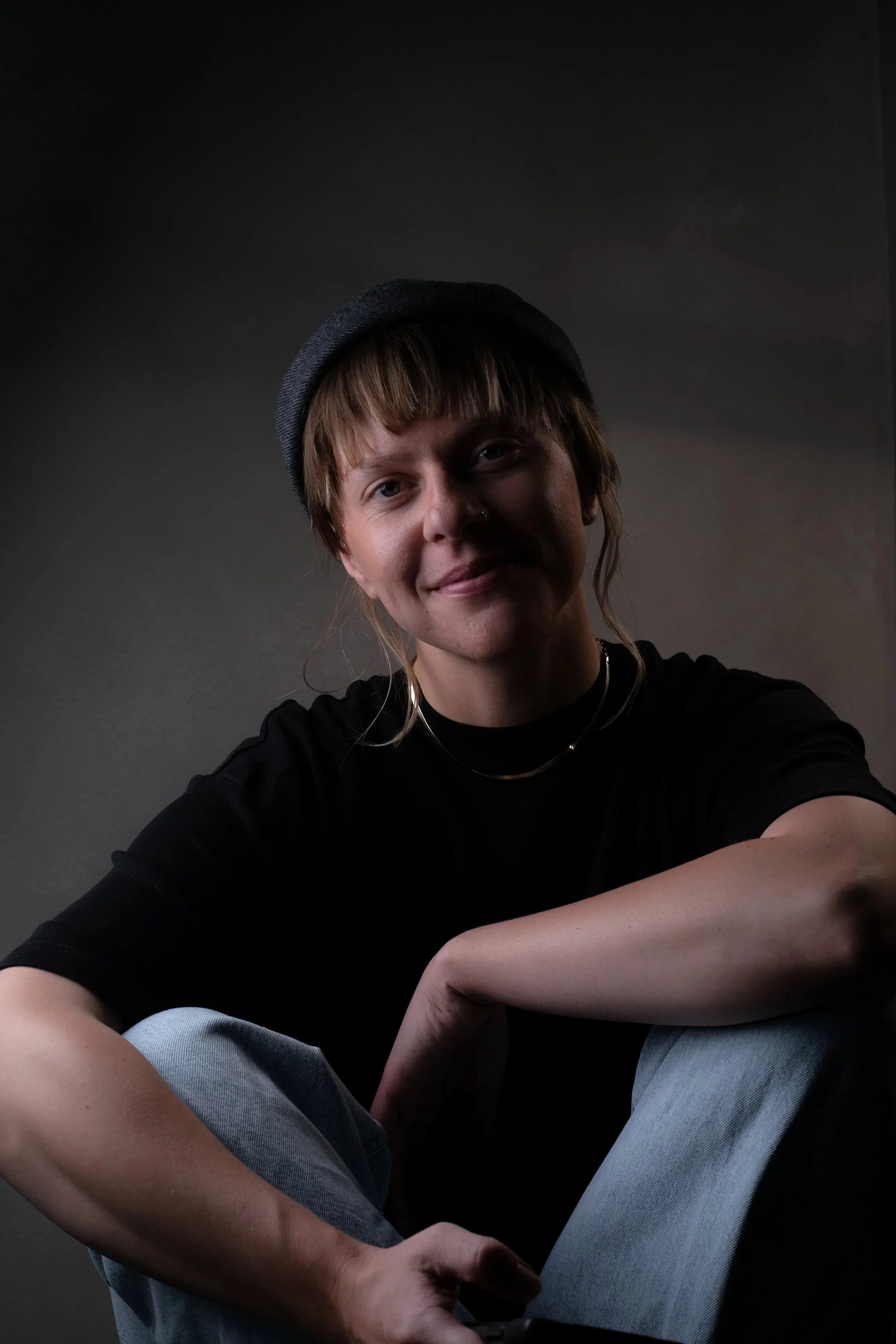 Portrait picture of Ola Getka, tattoo artist and choreographer based in Iceland wearing a gray beanie, black t-shirt, and layered necklaces, sitting against a dark background.