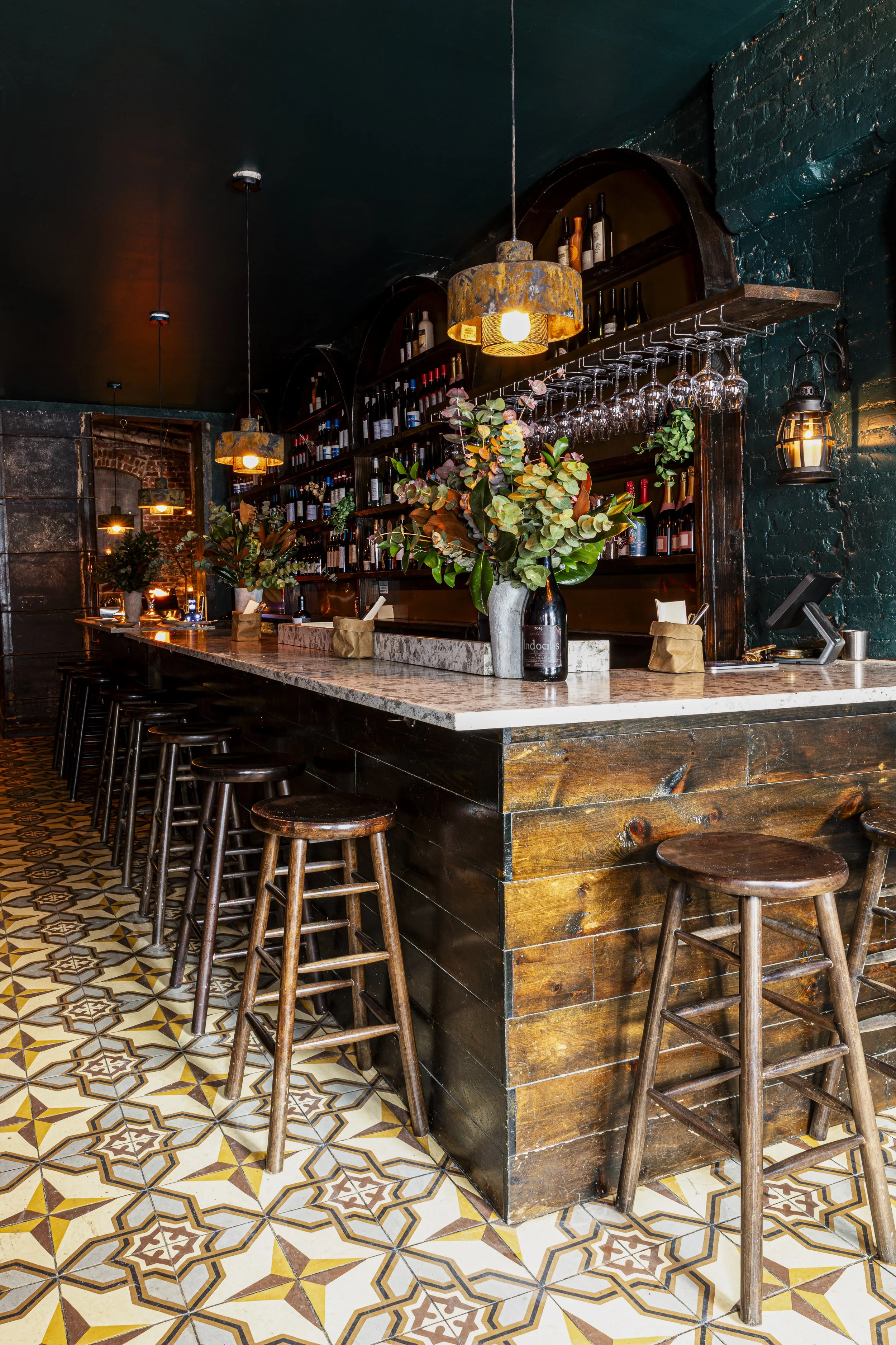 Interior of a cozy bar with wooden bar stools, a marble countertop, and a back wall filled with wine bottles and glasses, decorated with vases of green foliage and warm hanging lights.