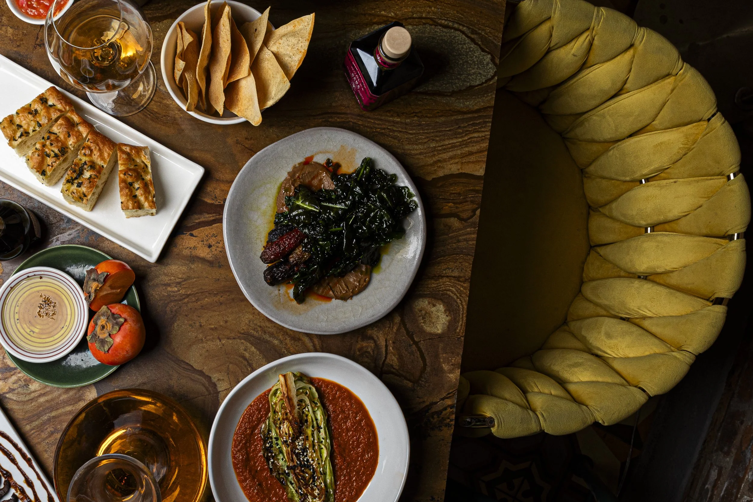 A top-down view of a dining table with various foods including a white rectangular plate of sliced bread topped with black sesame seeds, a bowl of pita chips, a wine glass with white wine, a white plate with a dark leafy vegetable dish, a small bowl of sauce, a white bowl with stuffed zucchini in tomato sauce, and a large yellow tufted chair.