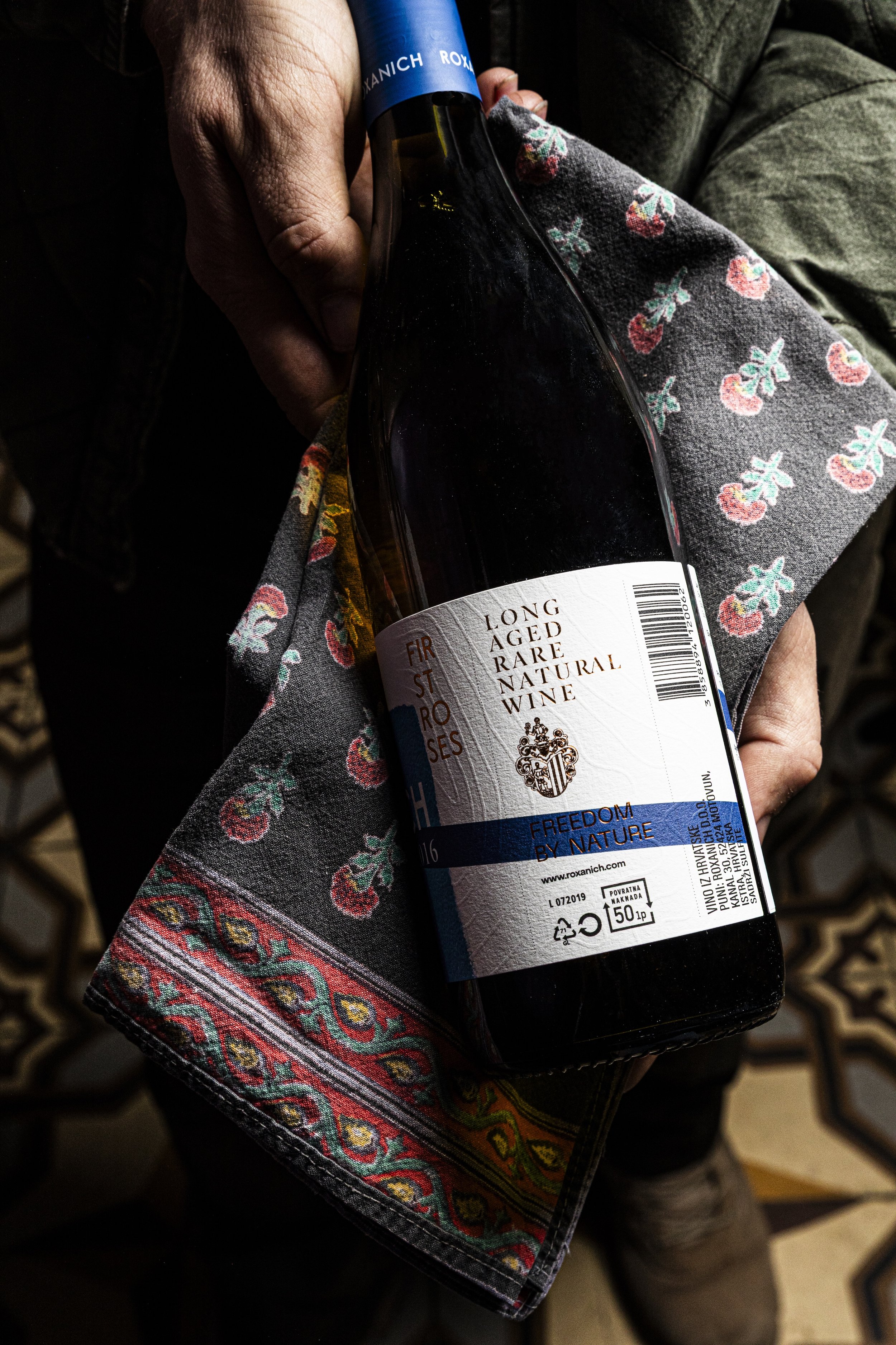 Person holding a bottle of long-aged, natural wine wrapped in a colorful, embroidered fabric with a pattern of cherries.