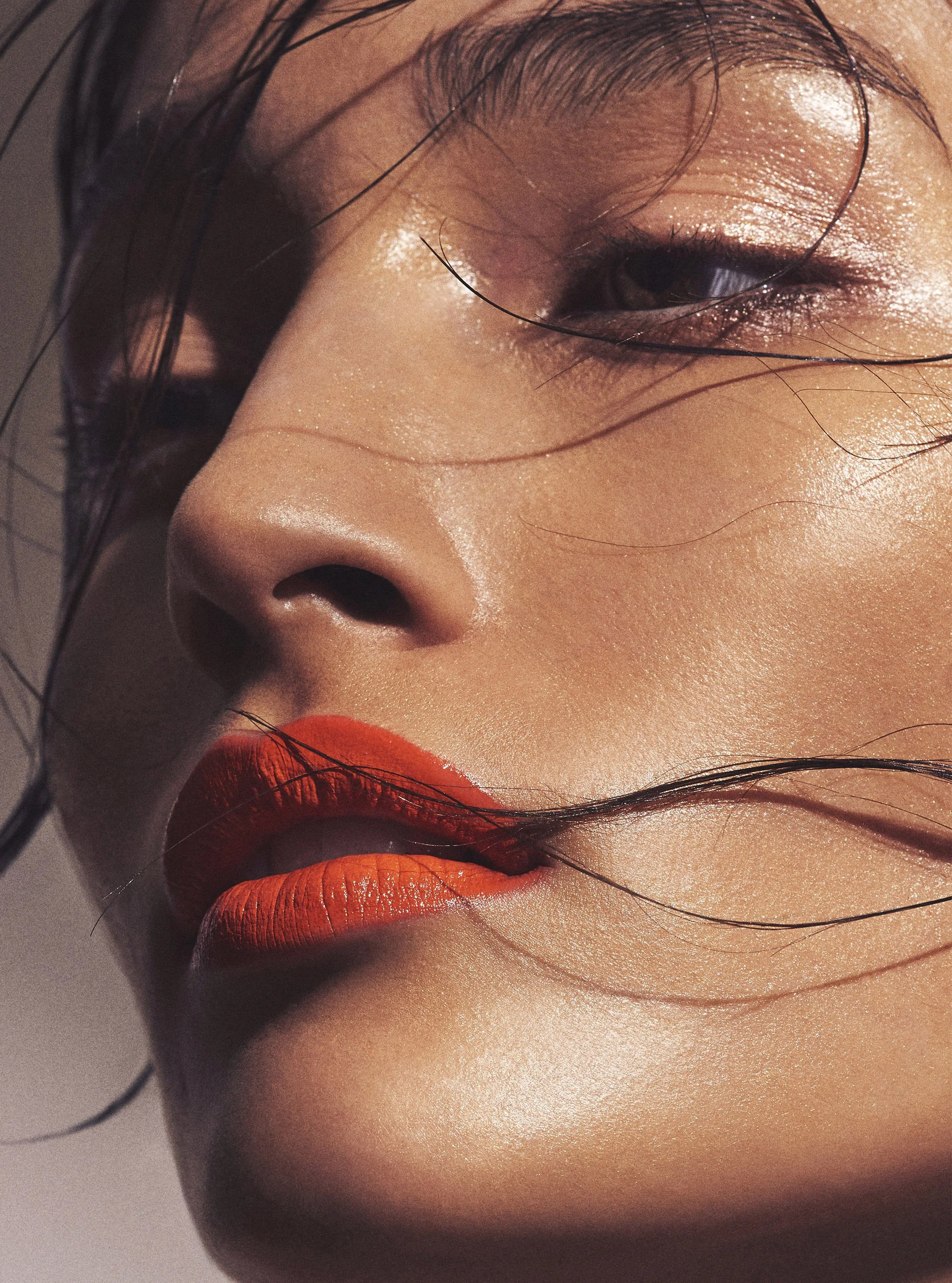 Close-up of a woman's face with shiny skin, orange lipstick, and wet hair strands.