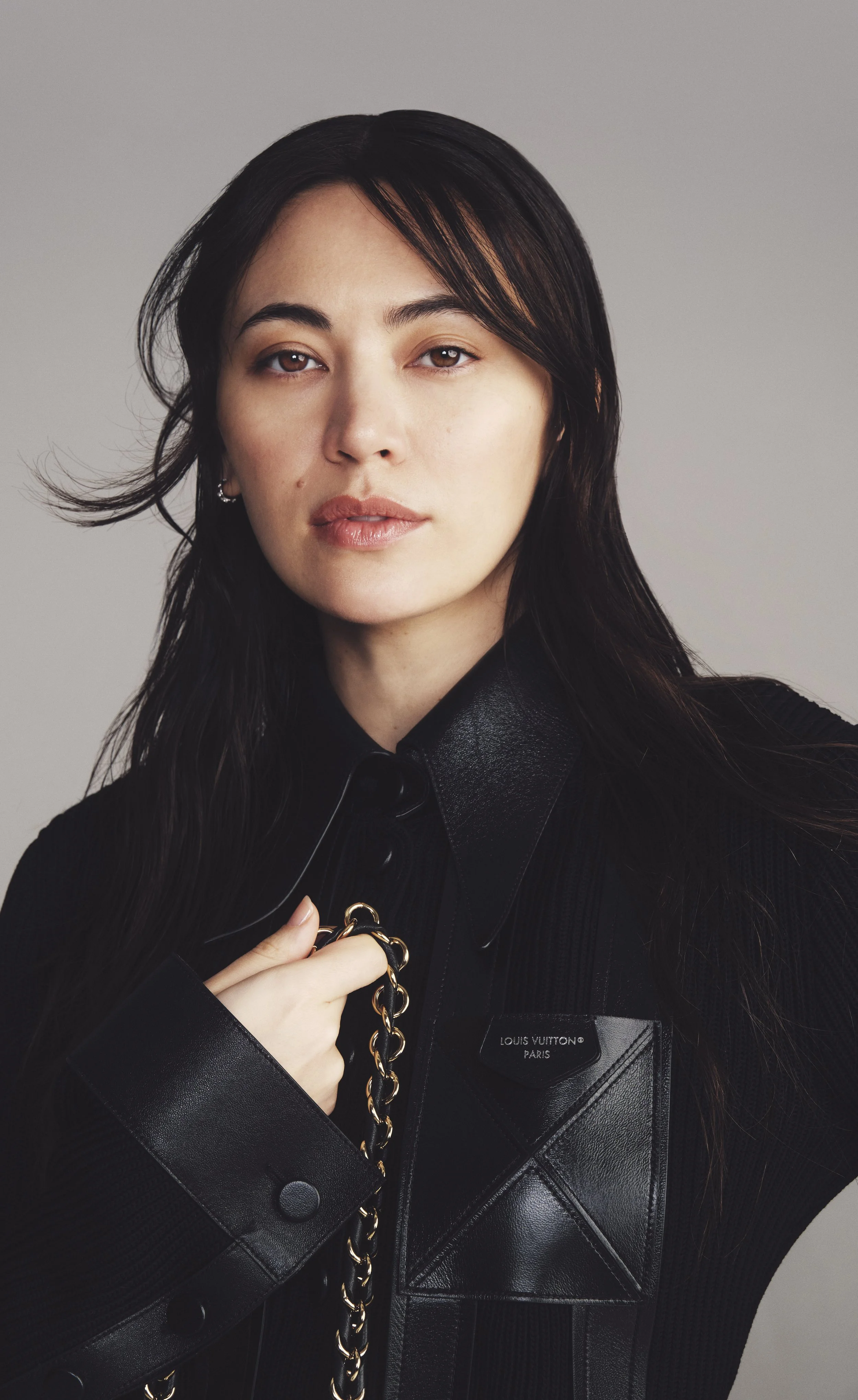 A woman with long black hair, wearing a black leather jacket with a Louis Vuitton Paris tag, holding a gold chain.