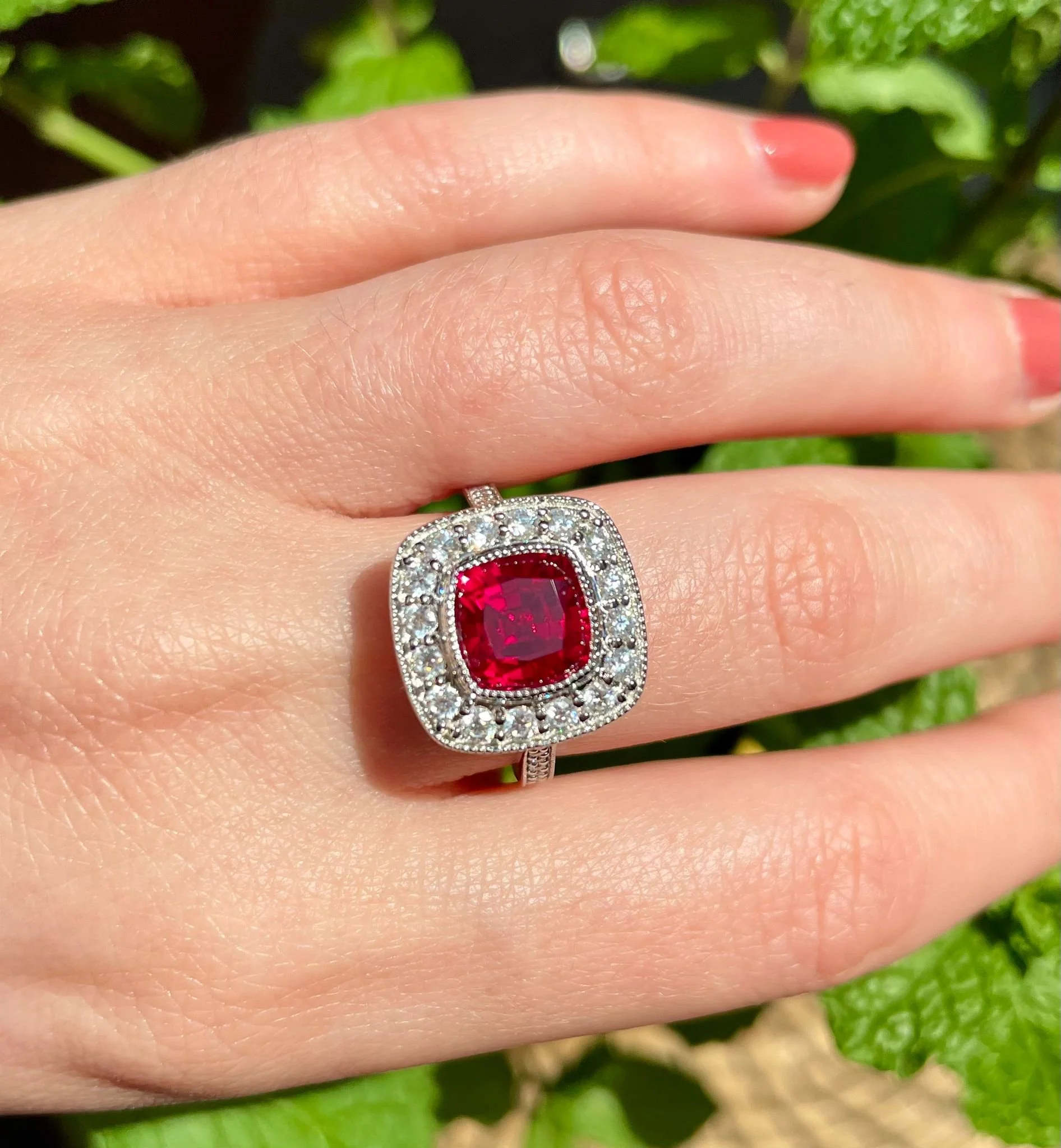 A hand with a silver ring featuring a large red gemstone surrounded by smaller clear gemstones, resting on green leaves.