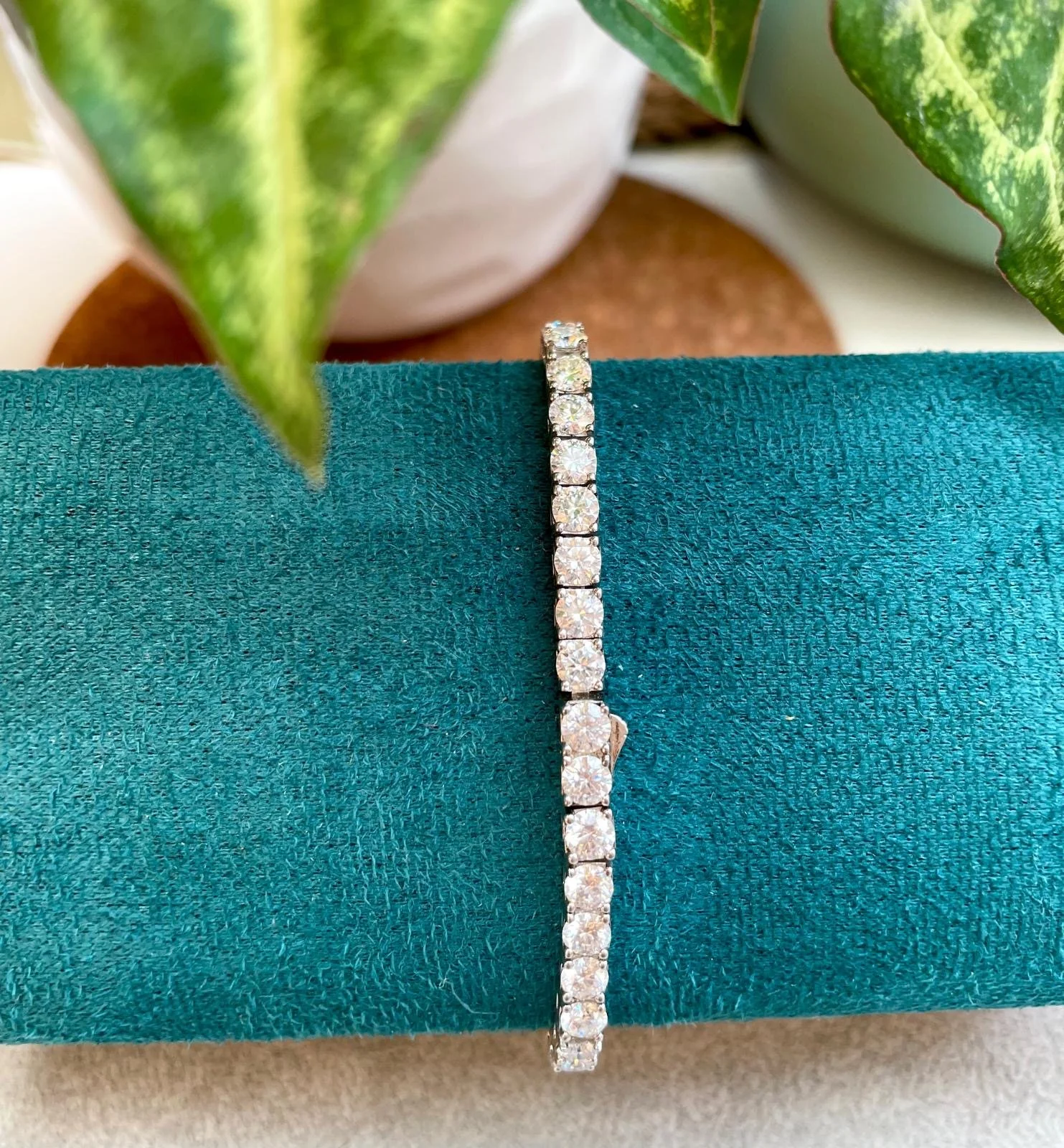 A silver bracelet with multiple round-cut diamonds displayed on a teal fabric background, with green leaves and potted plants in the background.
