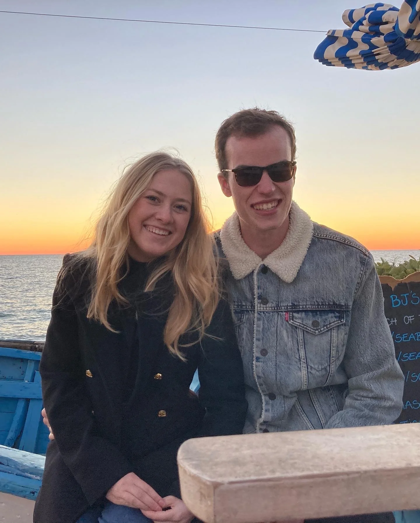 A young couple smiling at sunset near the water, with a blue and white striped umbrella overhead.