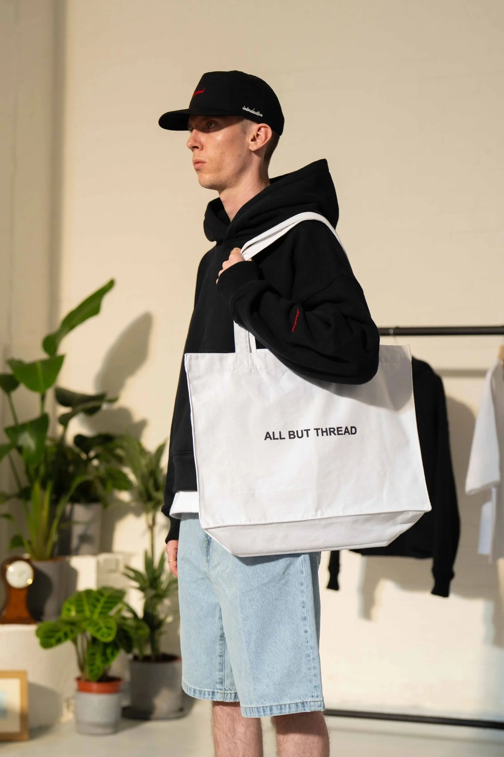 Young man wearing a black cap, black hoodie, and denim shorts, carrying a white tote bag with the text 'ALL BUT THREAD' in a room with potted plants and clothing displays.