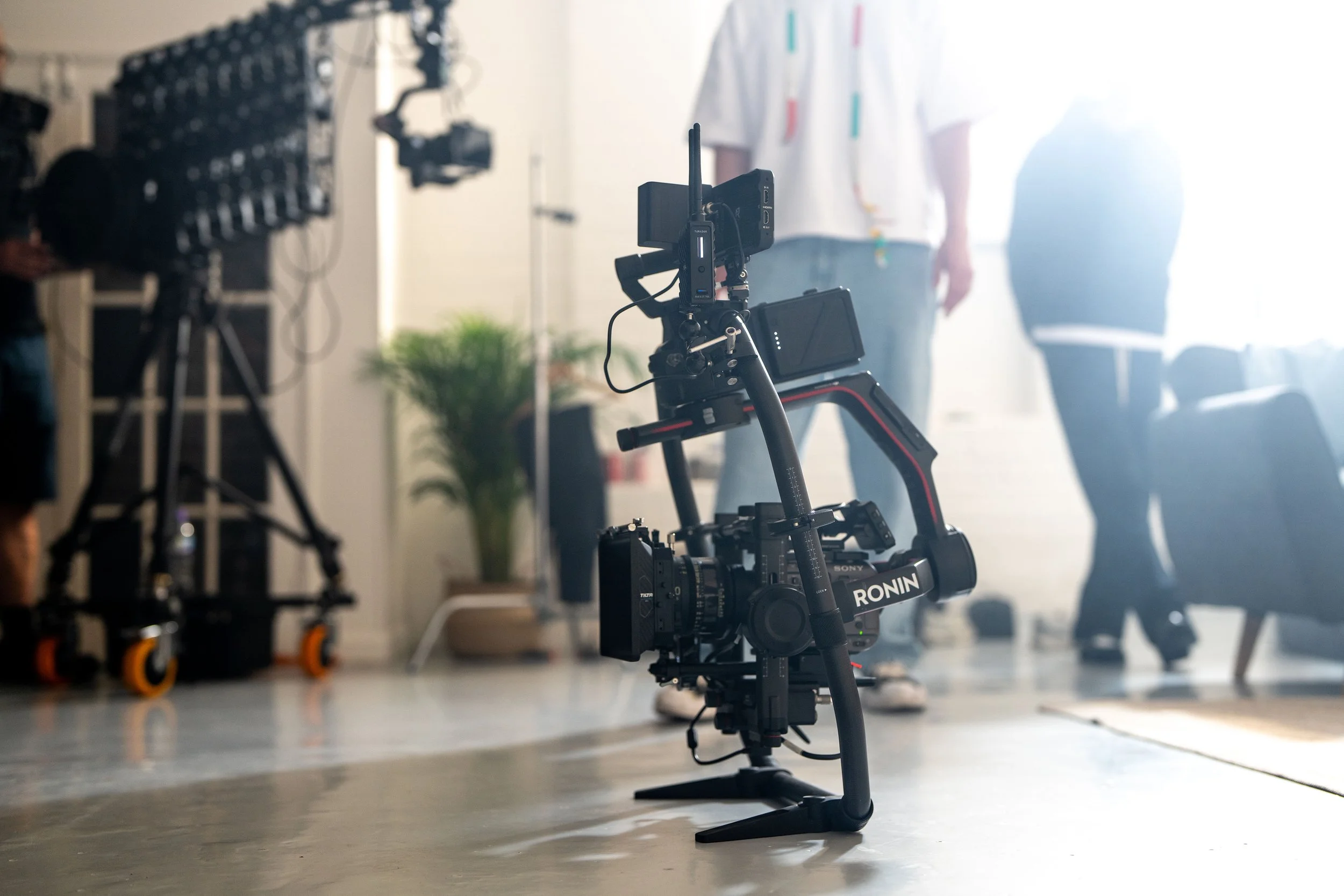 Camera mounted on a gimbal stabilizer in a studio setting, with people in the background and equipment around.