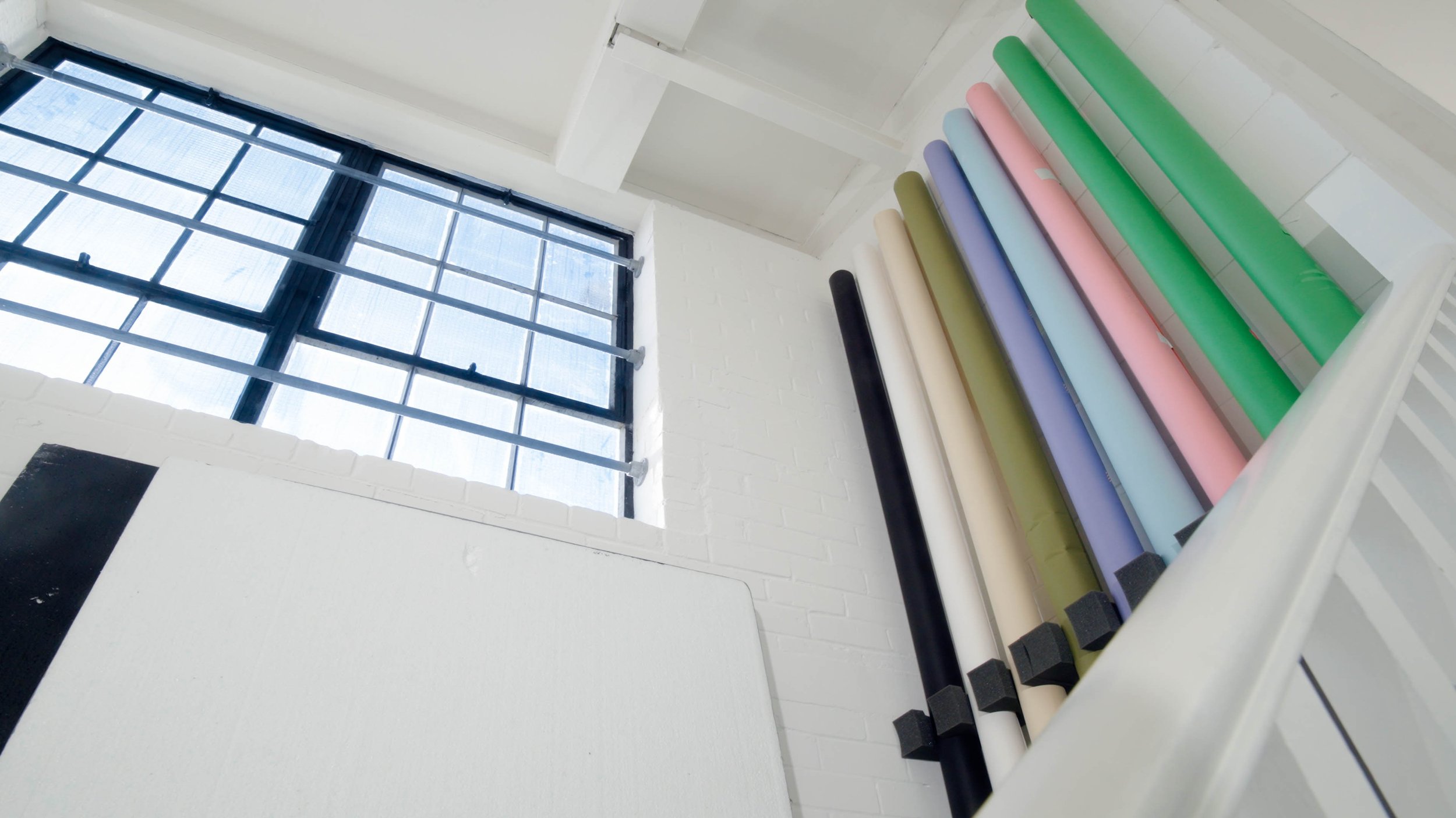 Colorful rolls of vinyl or paper stored on a white rack near a large window with metal bars, in a brightly lit room with white brick walls.