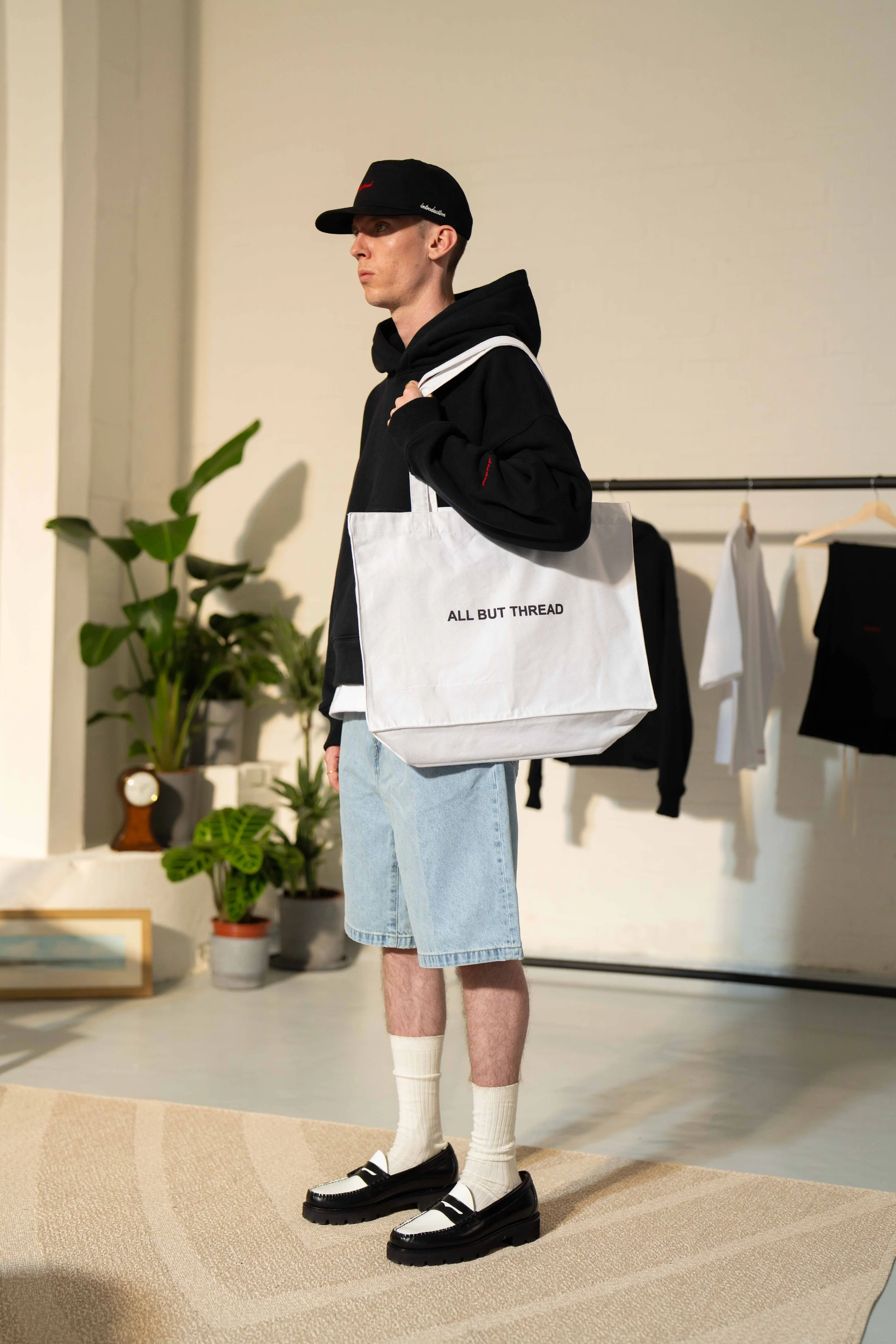A young man wearing a black baseball cap, black hooded sweatshirt, light denim shorts, white socks, and black and white loafers, standing indoors on a beige rug with a white tote bag that reads 'ALL BUT THREAD'.