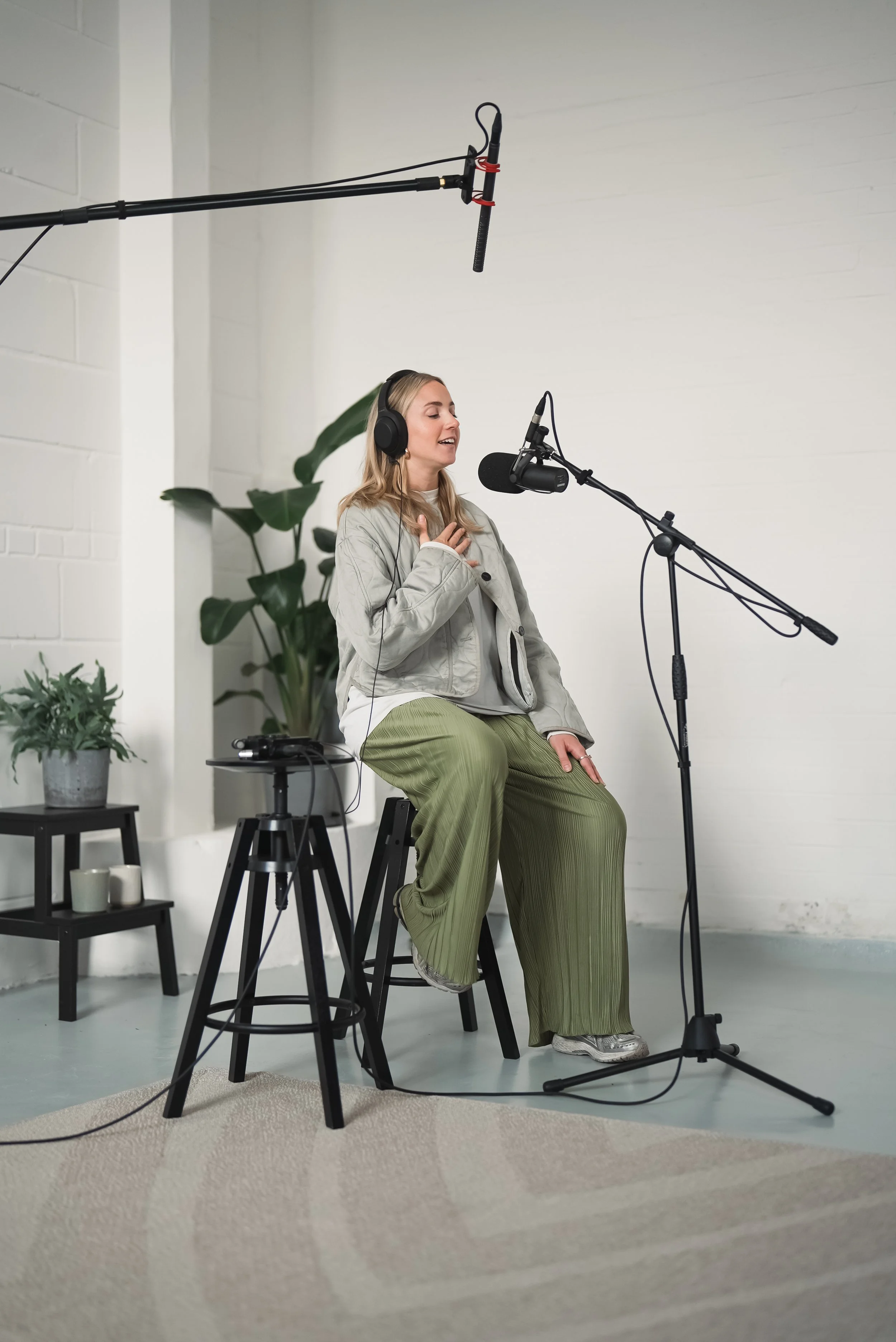 A woman with blonde hair wearing headphones, a light gray jacket, green wide-leg pants, and white sneakers, singing into a microphone in a recording studio with a white wall, green plants, and a small black table.
