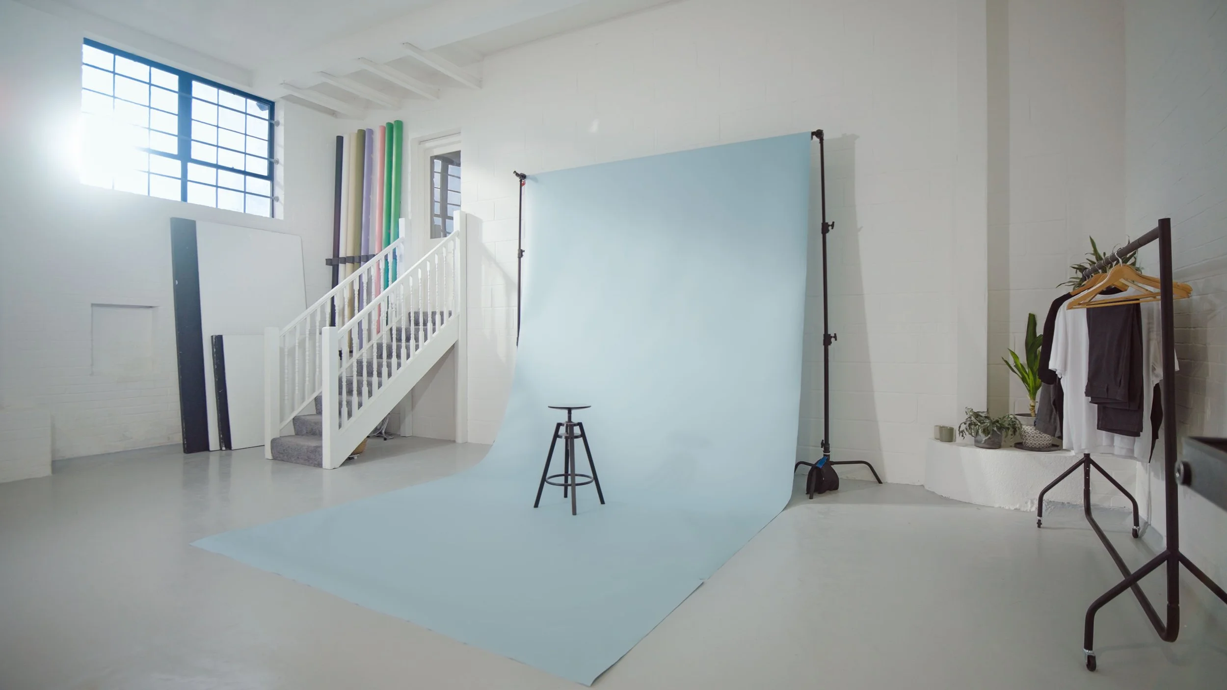 Photo of a photography studio with a blue backdrop, stool, clothing rack, and potted plants, illuminated by natural light from windows.