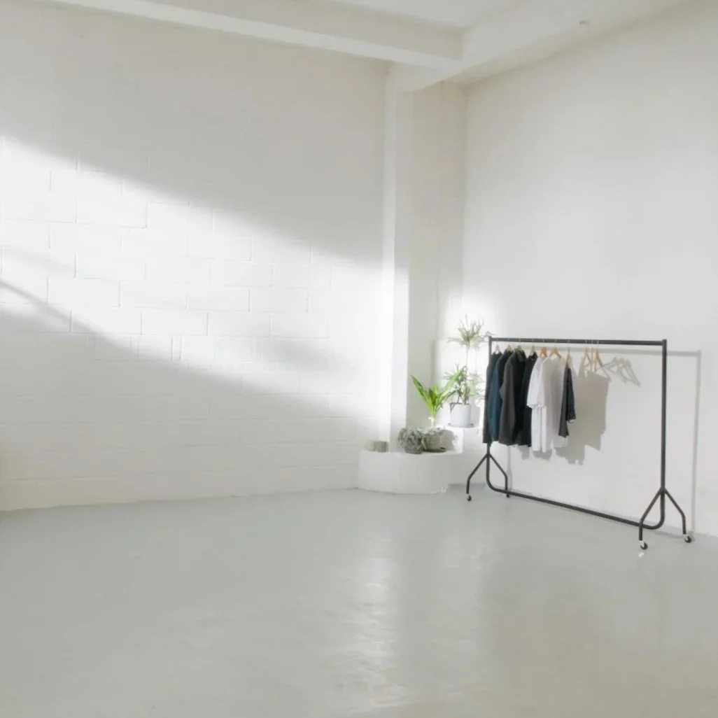 Minimalist white room with a clothing rack holding a few shirts and jackets, a small green plant, and sunlight casting shadows on the wall.