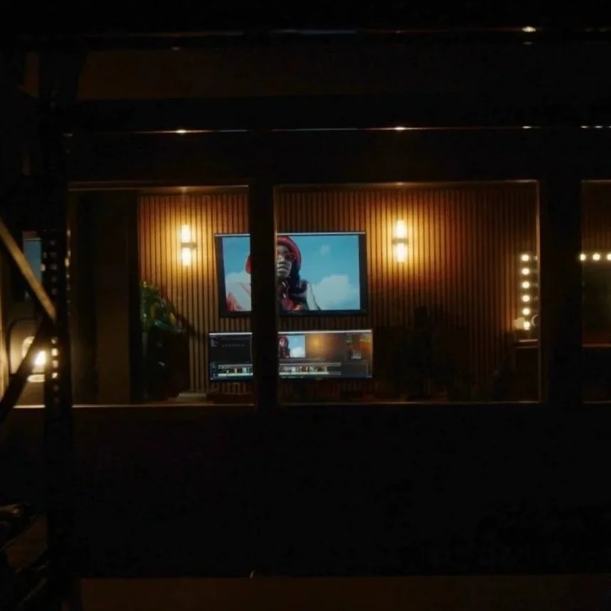 A viewing room with two large monitors showing a person wearing headphones against a cloudy sky background, with warm lighting and a wooden wall.