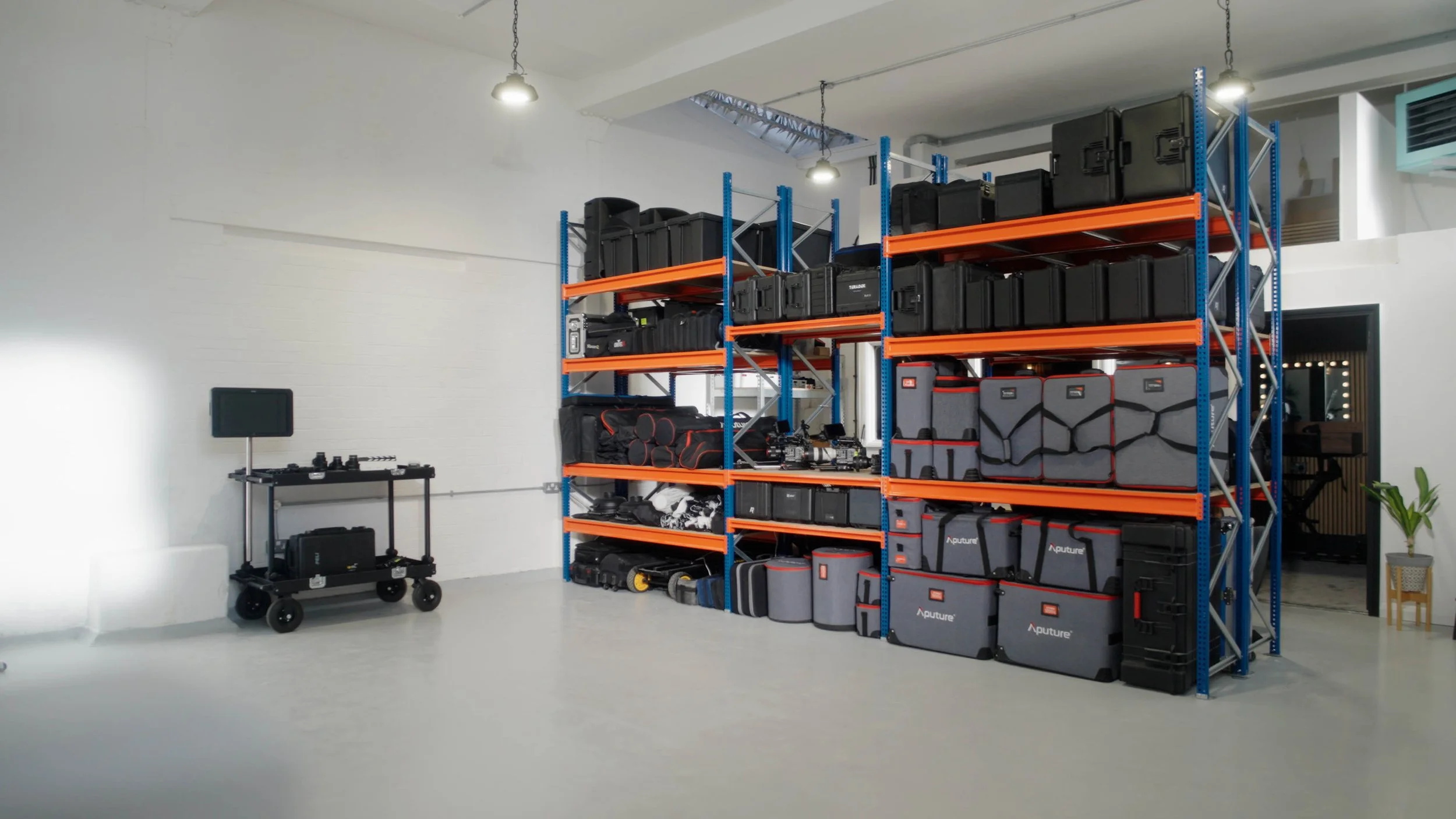 A storage room with large orange and blue metal shelving filled with black and gray equipment cases. There is a small wheeled cart with electronic equipment and a monitor on the left side of the room, and a potted plant on a small stand near a door on the right side.