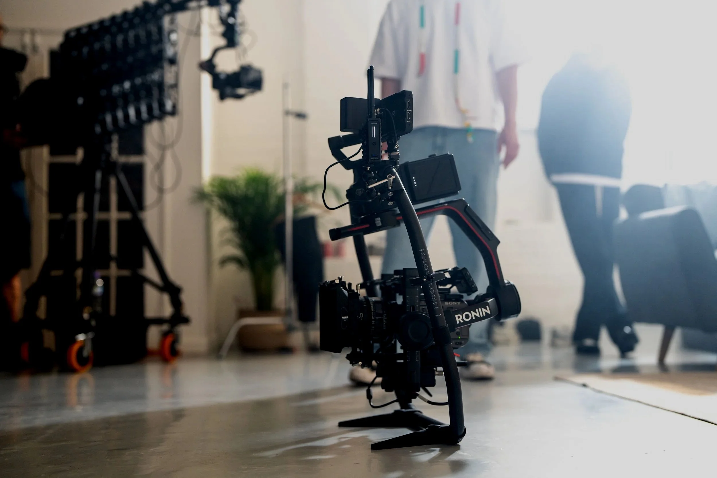 Photography camera mounted on a gimbal stabilizer in a studio with blurred people and furniture in the background.