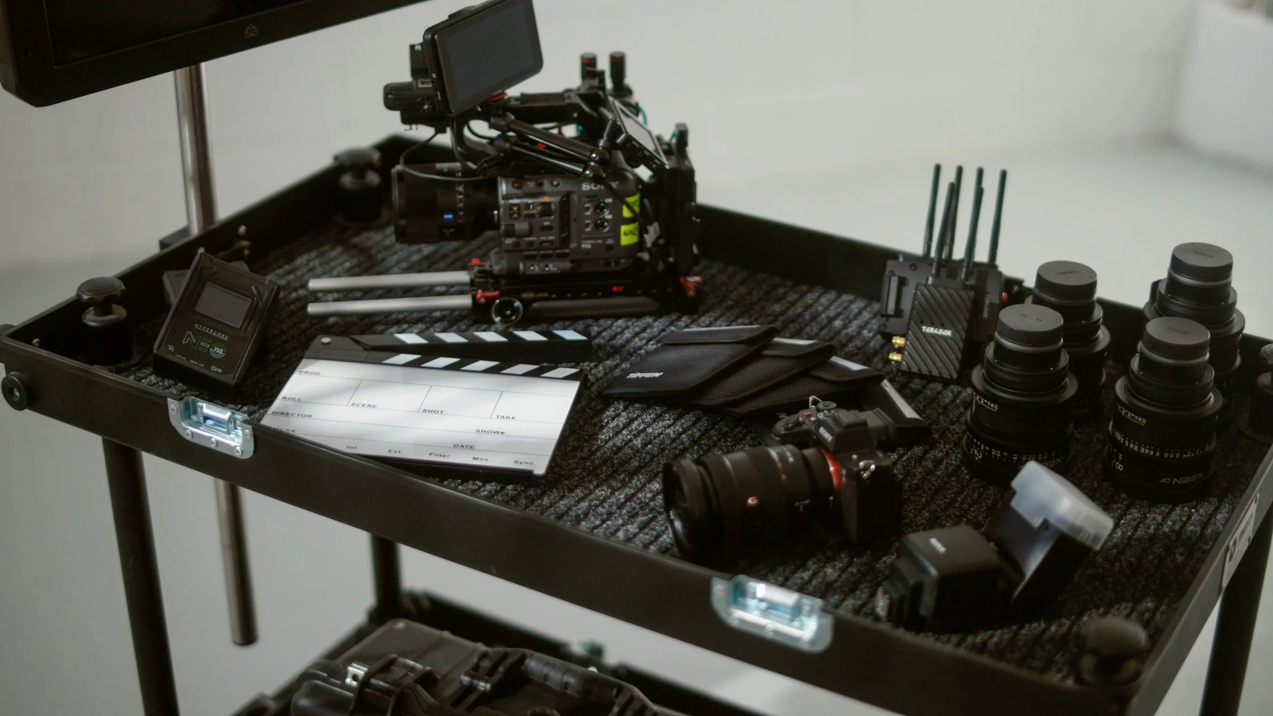 Film production equipment on a black table, including a camera, lenses, monitors, batteries, a clapperboard, wireless transmitters, and accessories.