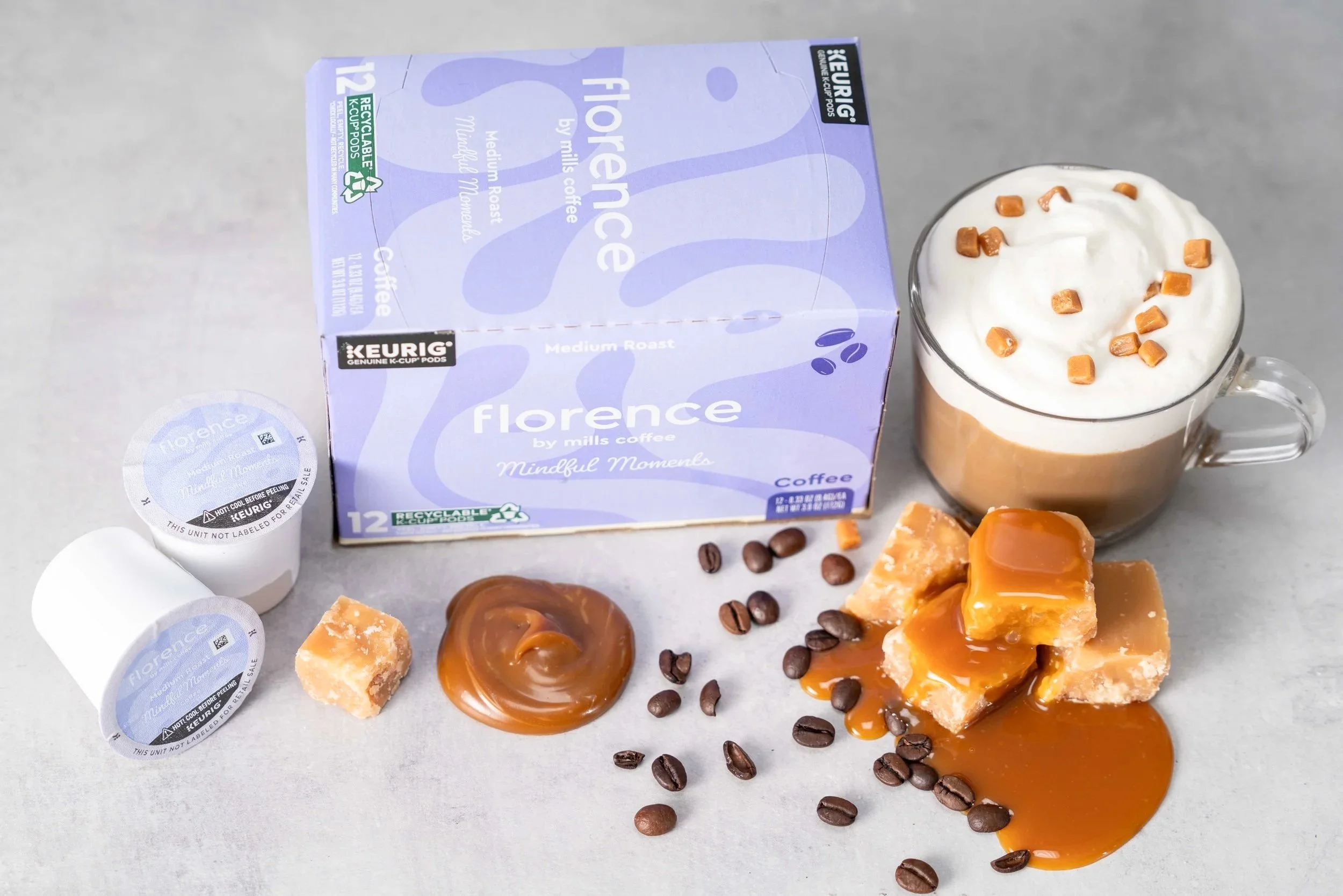 A box of Florence medium roast coffee pods, two caramel coffee drinks topped with whipped cream and caramel candies, scattered caramel candies, coffee beans, and caramel sauce on a light surface.