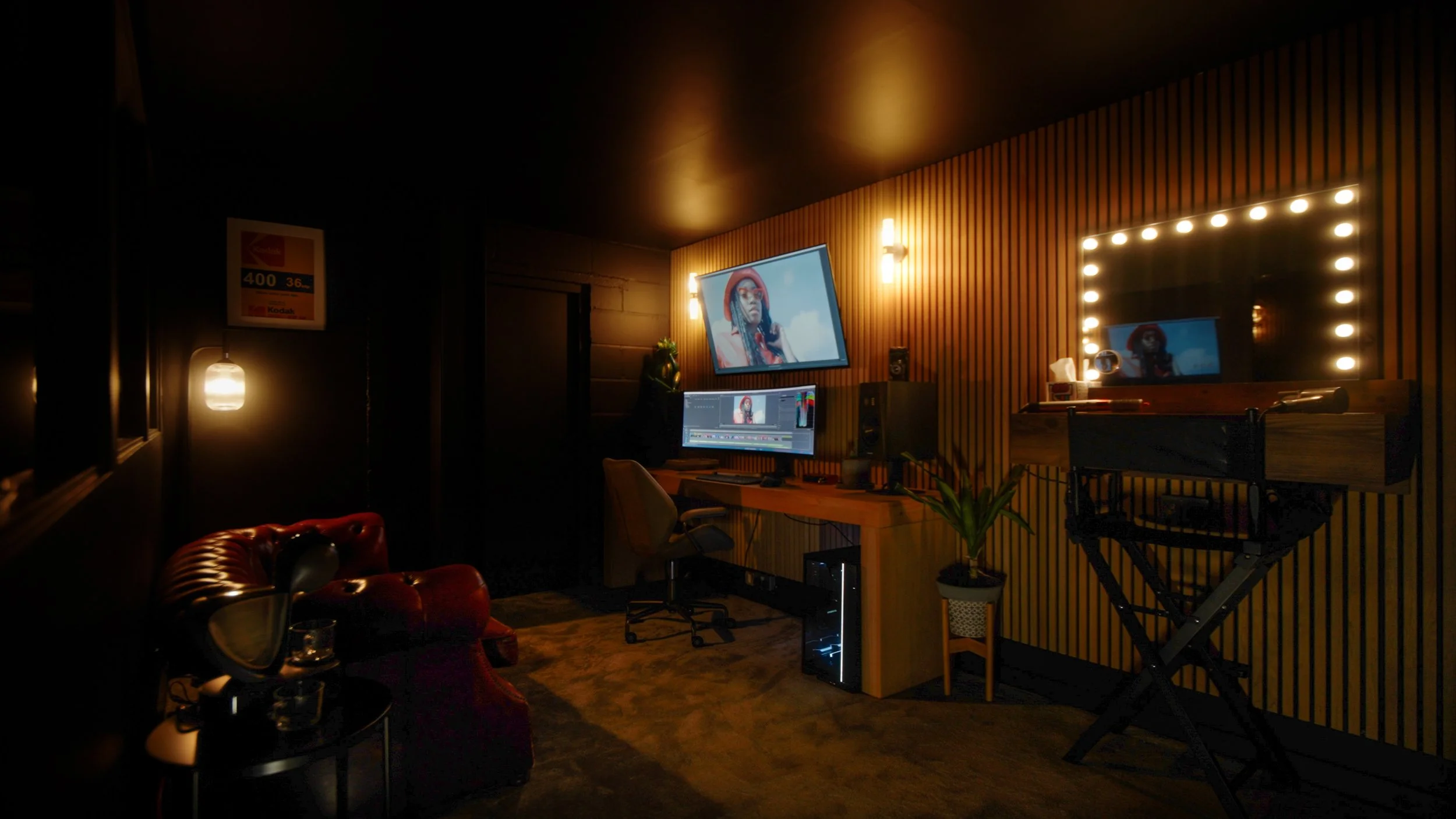 A dimly lit music or recording studio room with a large illuminated mirror, a computer setup on a wooden desk, a leather armchair, a red leather couch, and a film or photo shoot light. The room has wood paneling on one wall and a dark ceiling.
