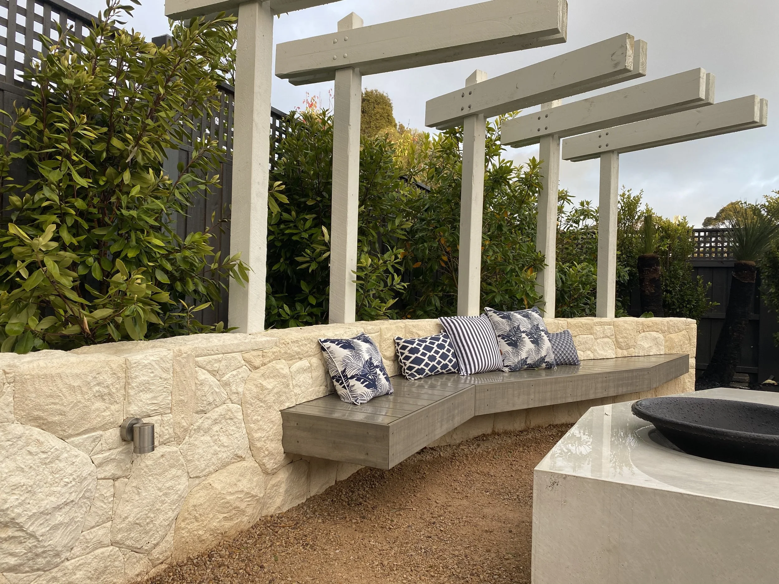 The Block 2021, Exotic Living. Outdoor seating area with a stone and wood bench, decorative pillows, and lush green plants.