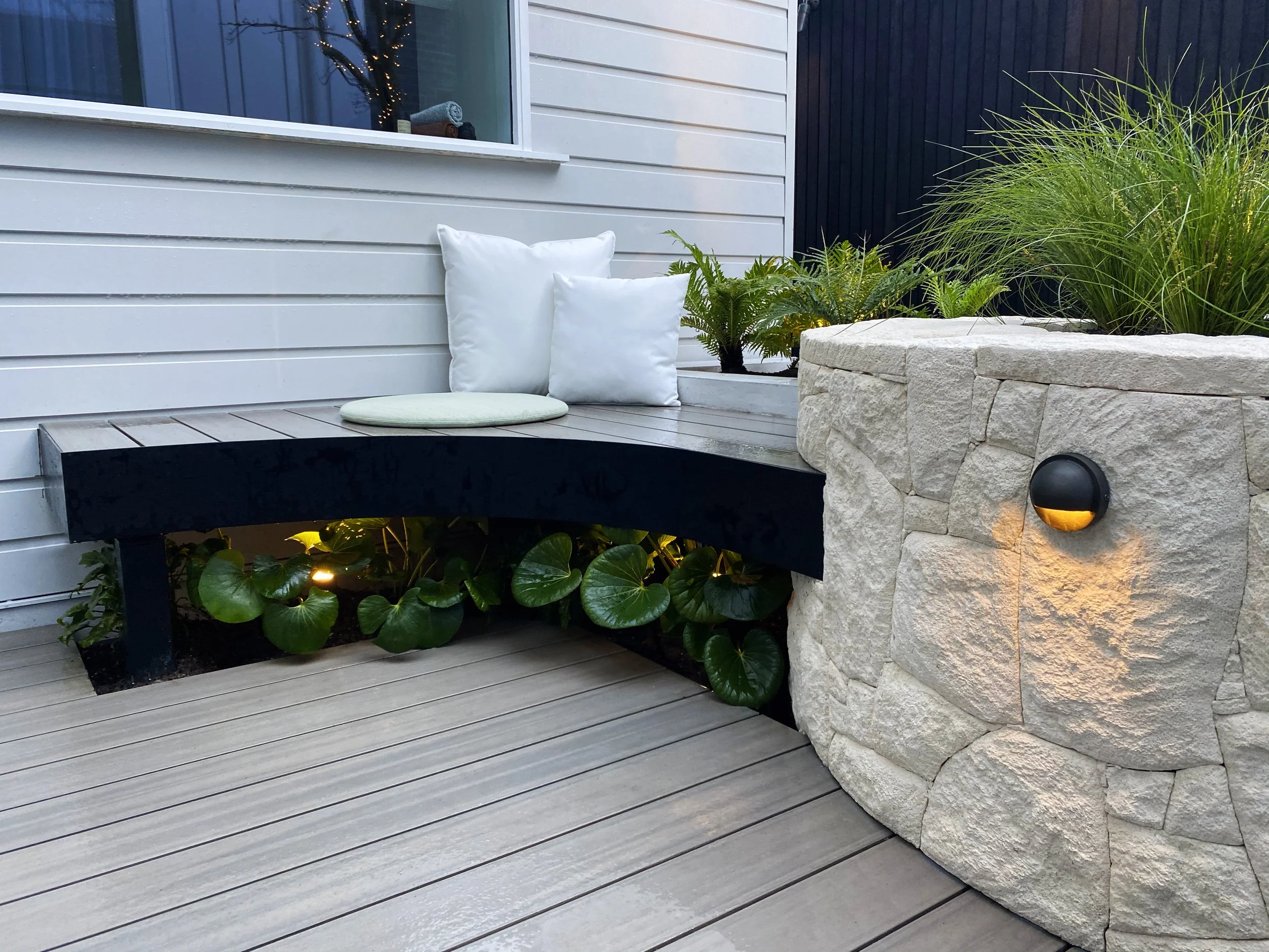 The Block 2020, Exotic Living. Outdoor courtyard with a black curved bench with white pillows and green planting underneath, a large stone planter with greenery, and a black wall in the background.