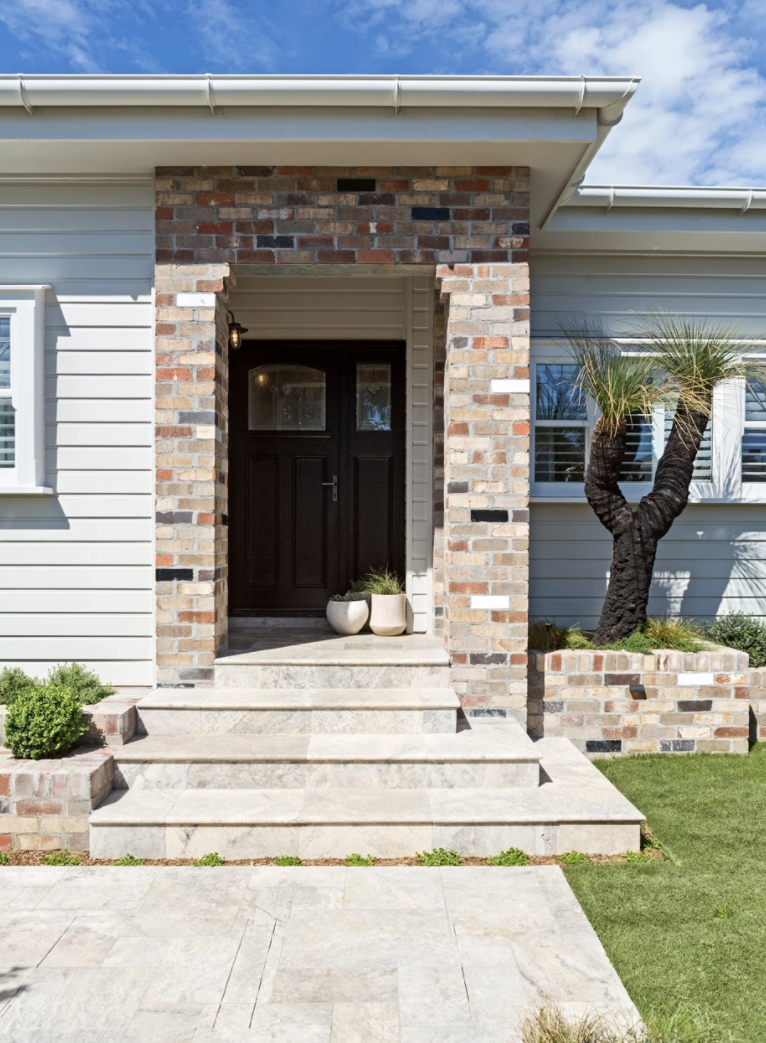 The Block 2020, Exotic Living. Front entrance of a modern house with a brick frame around the door, black front door, and steps leading up to the door. There is a small landscaped area with a twisted tree and potted plants beside the entrance.