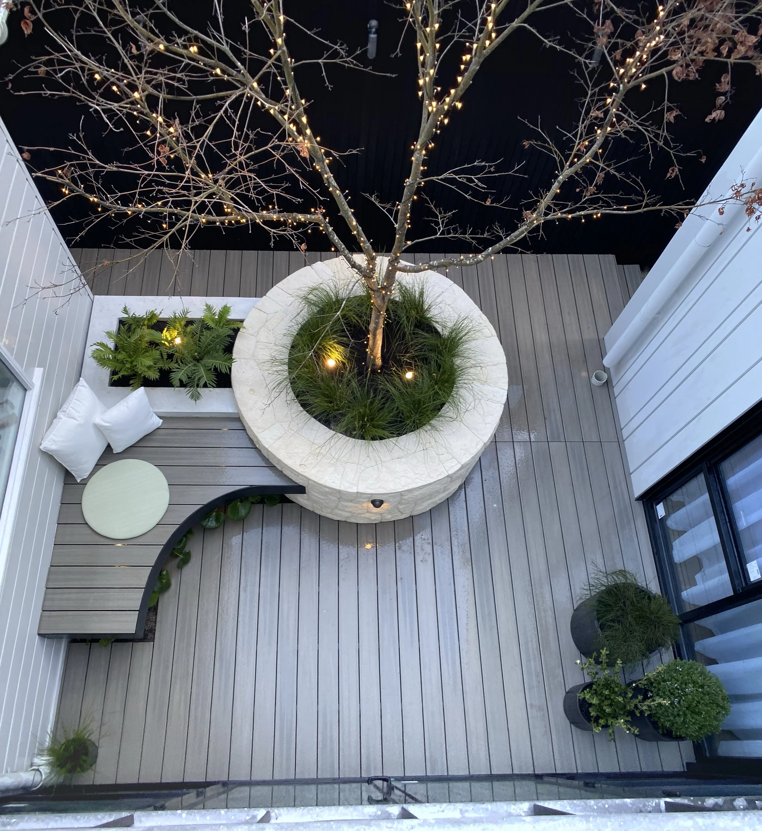 The Block 2020, Exotic Living. View of a small outdoor courtyard with wooden decking, a white curved bench with white pillows, potted plants, and a large tree with string lights wrapped around its branches.