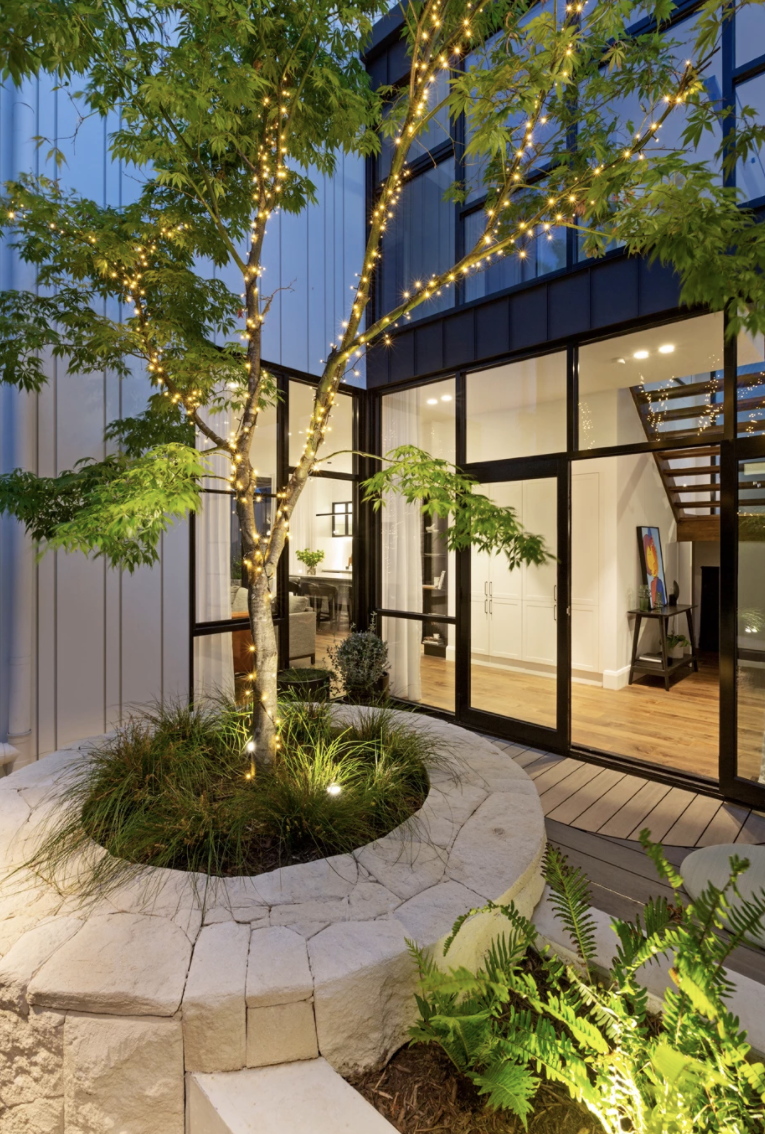 The Block 2020, Exotic Living. A modern house with large glass doors and windows, a tree wrapped with string lights, and landscaped outdoor space with greenery and a stone border.