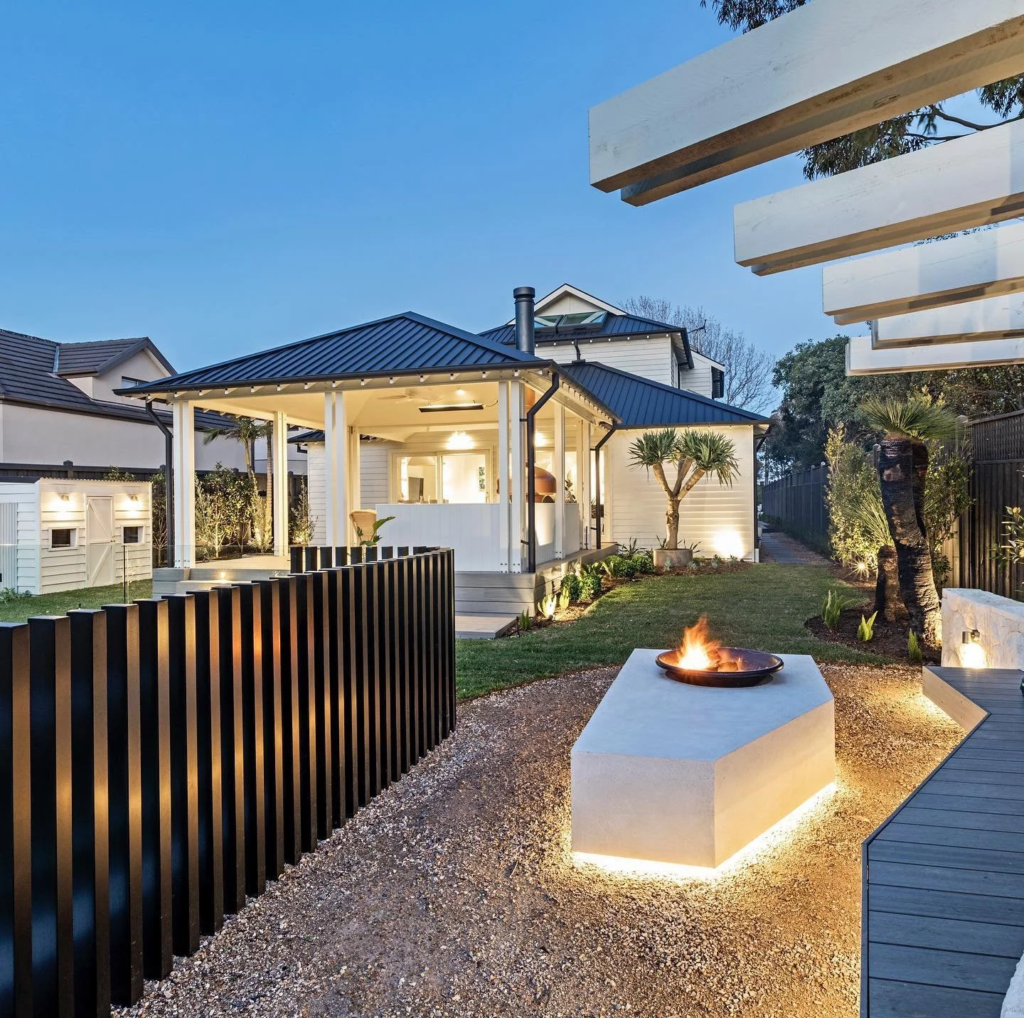 The Block 2021, Exotic Living. Modern backyard with fire pit, black metal fence, and covered patio, house with lit interior, sunset sky, and landscaped garden with trees and plants.