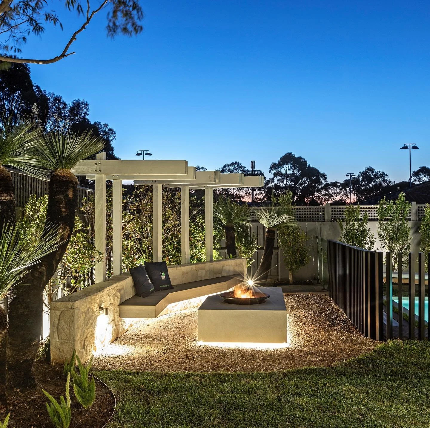 A modern backyard patio at dusk with a built-in stone bench, a fire pit with a glowing fire, and tropical plants including palm trees. In the background, there is a swimming pool and outdoor lighting, with a dark fence and trees against a twilight sk