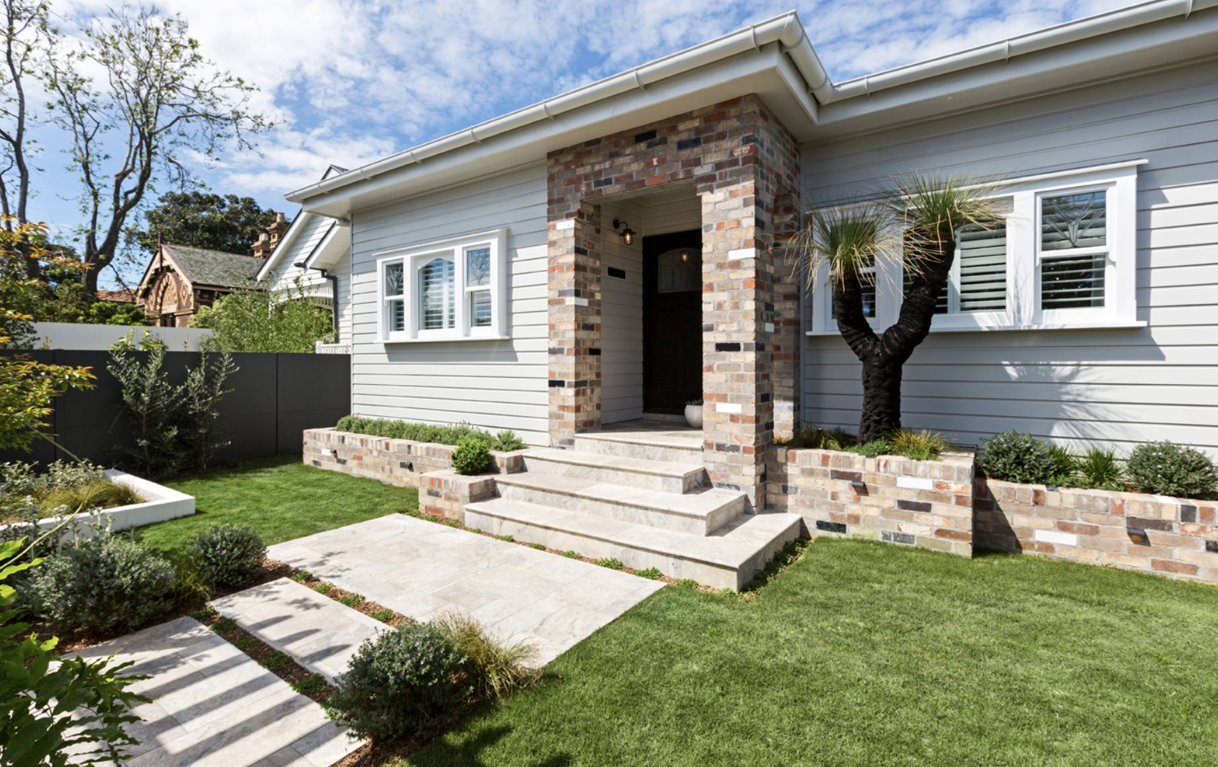 The Block 2020, Exotic Living. A suburban house with a brick and light gray siding exterior, front porch with steps, a landscaped yard with grass and plants, and a tree near the house.
