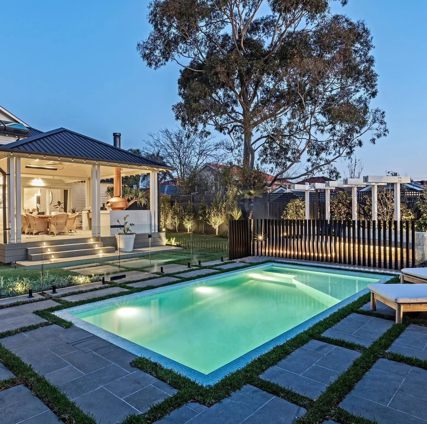 The Block 2021, Exotic Living. Backyard with a lit swimming pool, patio area with lounge chairs, a house with a covered porch, and tall trees at dusk.