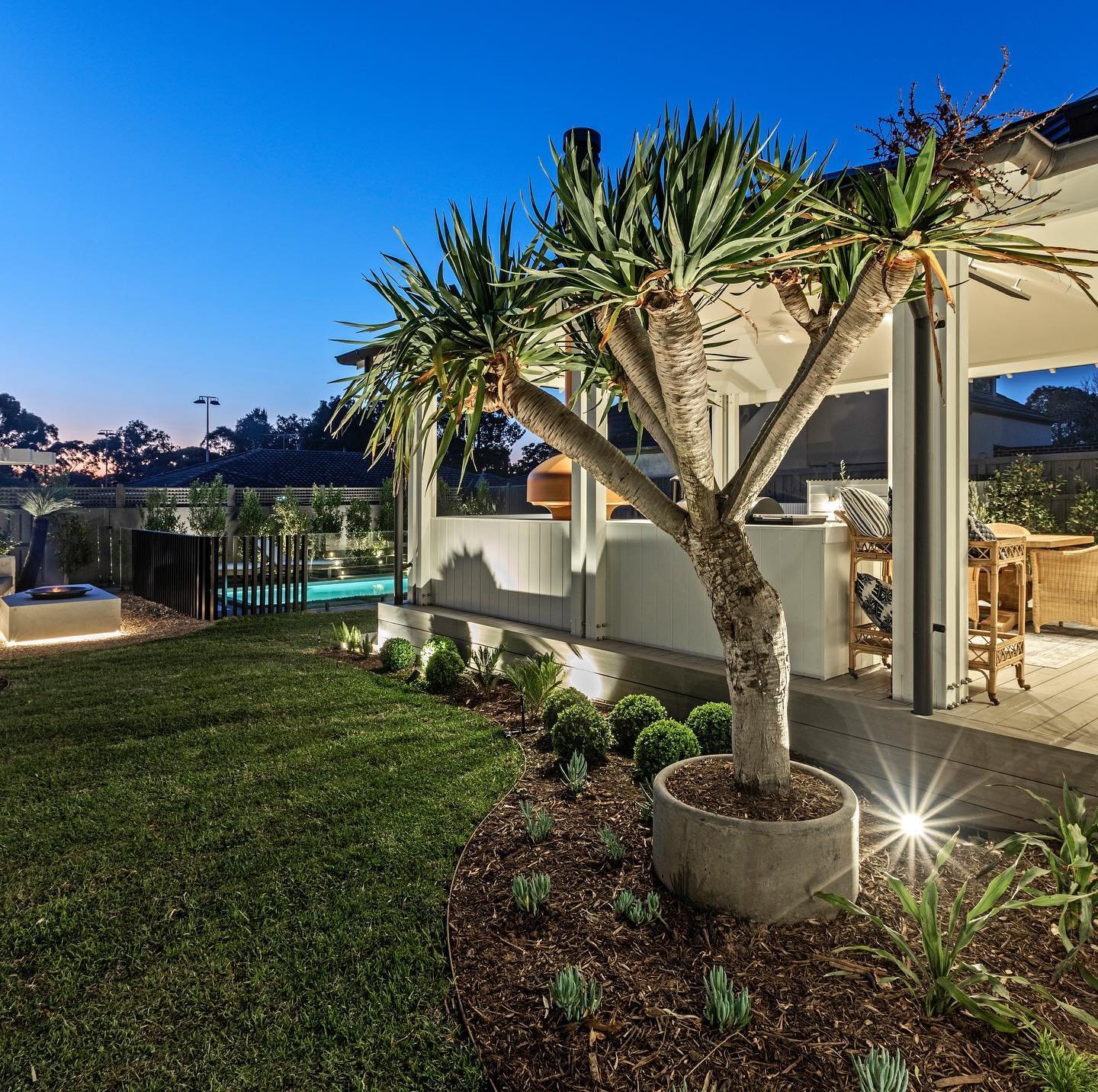 The Block 2021, Exotic Living. A backyard at dusk with a large potted tropical tree, well-manicured lawn, outdoor living space with wicker furniture, and a pool in the background under a darkening sky.