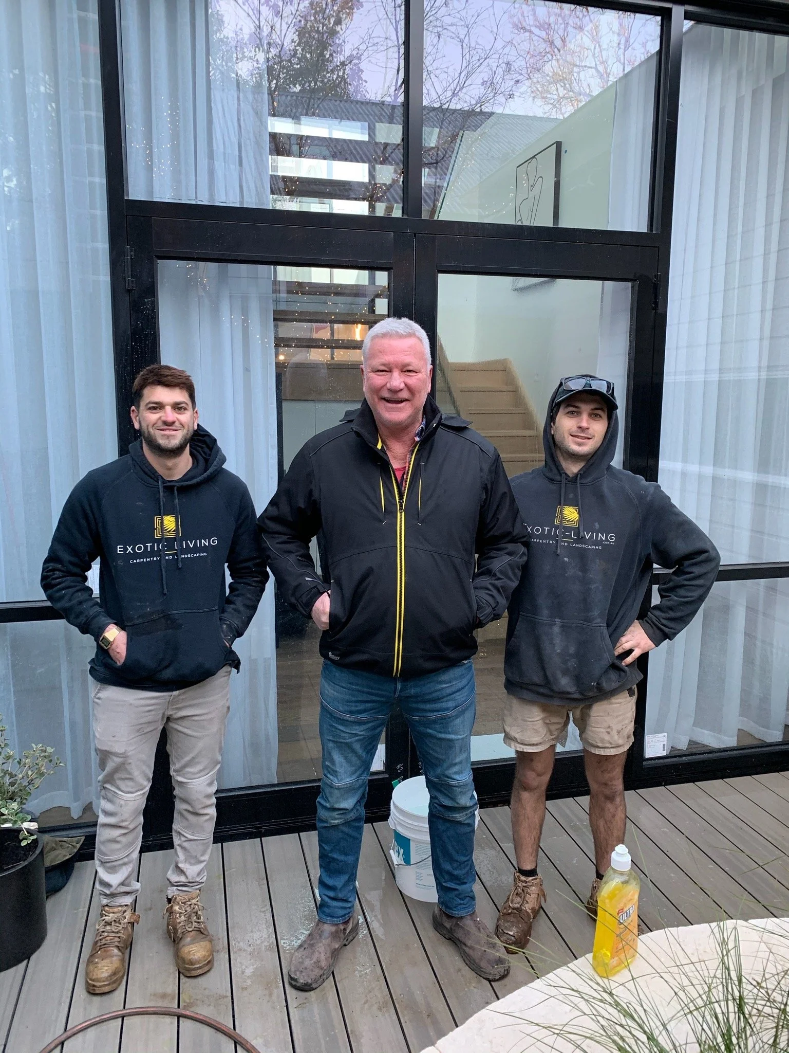 The Block 2020, Exotic Living. Three men standing outside on a wooden deck in front of large glass doors and windows, smiling. The two younger men are wearing black hoodies with a yellow logo, and the Scotty Cam is wearing a black jacket.