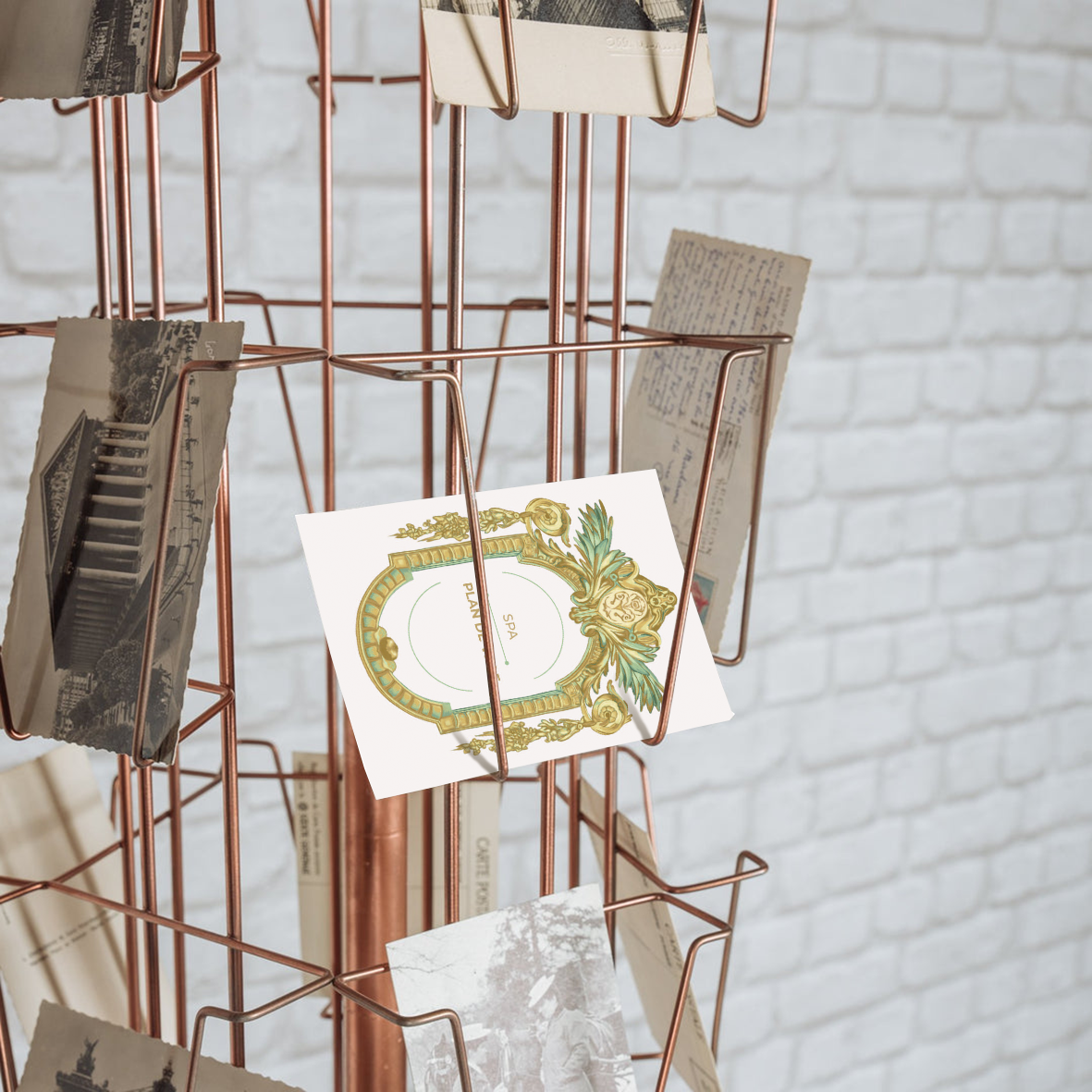 Un porte-affiches en fil métallique rose doré avec des photographies anciennes accrochées, situé devant un mur en briques blanches.