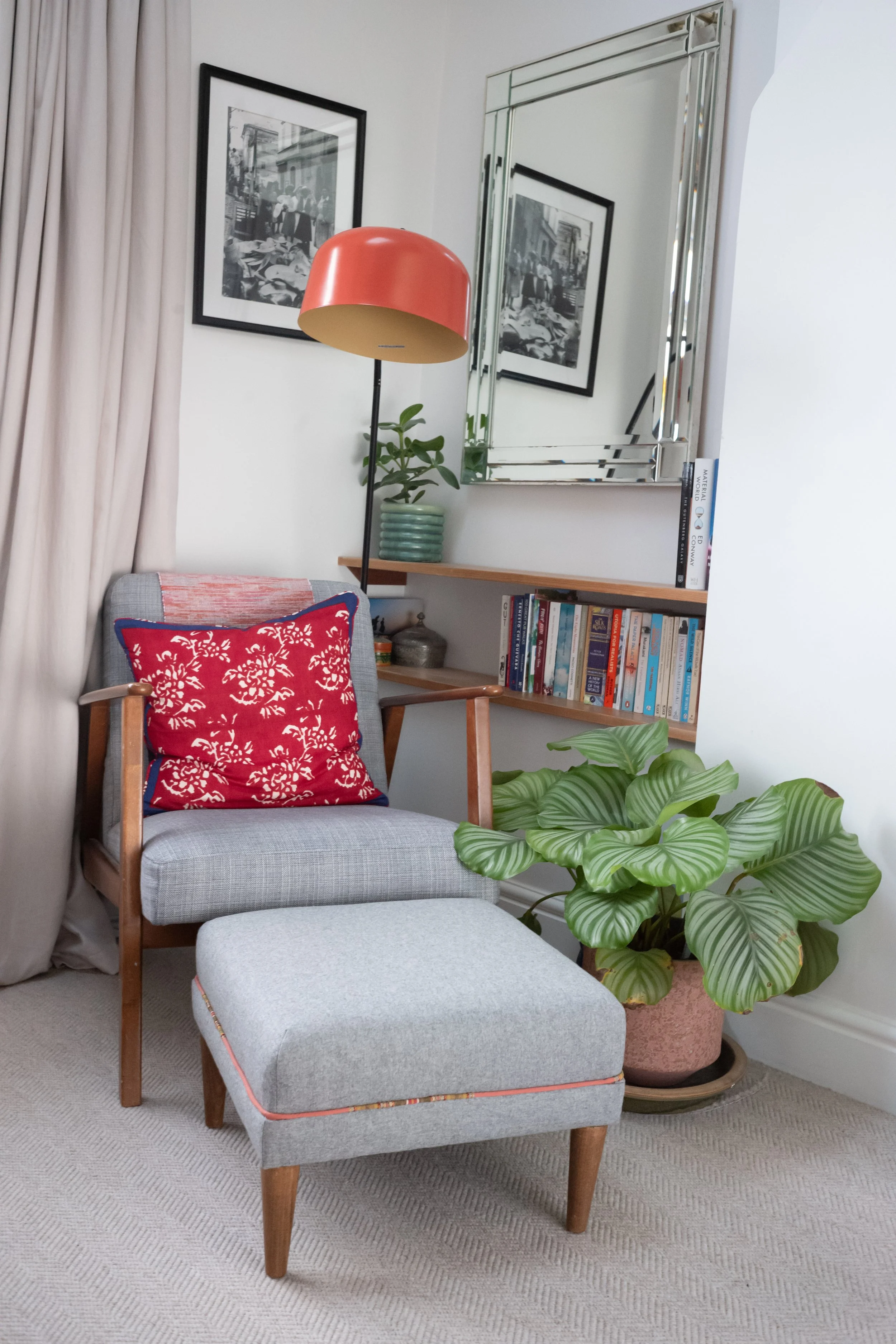 A cozy corner of a living room with a gray armchair and matching ottoman, a red decorative pillow, a large leafy plant in a pink pot, a wooden shelf with books, a green potted plant, a mirror, and black-and-white framed photographs on the wall.