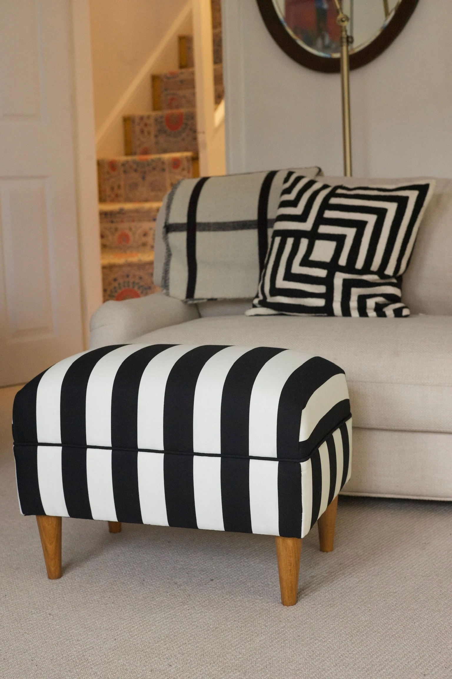 Black and white striped ottoman in front of a beige sofa with a black and white patterned pillow and draped blanket, against a background of a staircase with a colorful runner.