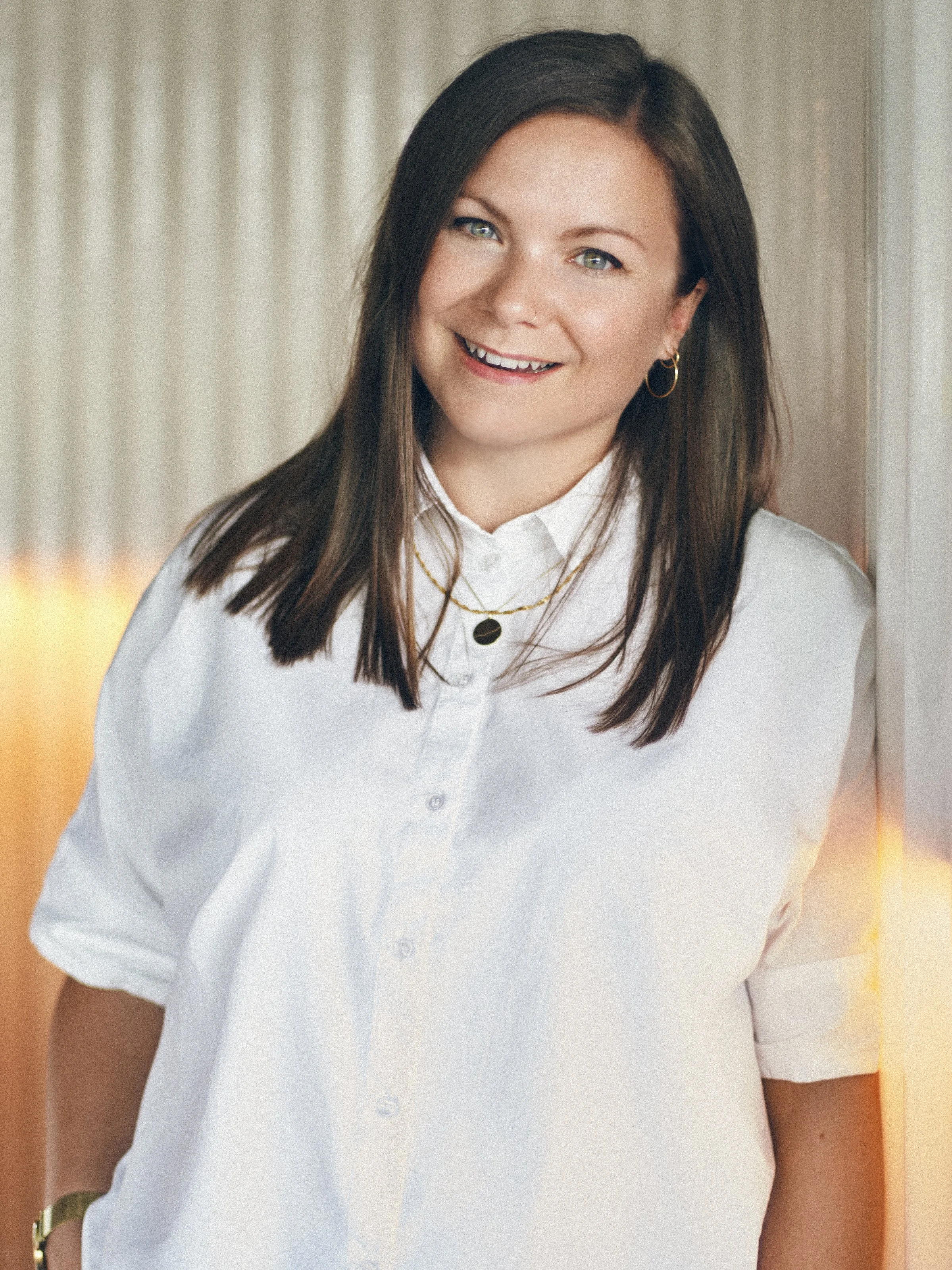 A woman with dark brown hair and blue eyes, smiling, standing indoors near a window with beige curtains, wearing a white button-up shirt with rolled sleeves, gold hoop earrings, and layered necklaces.