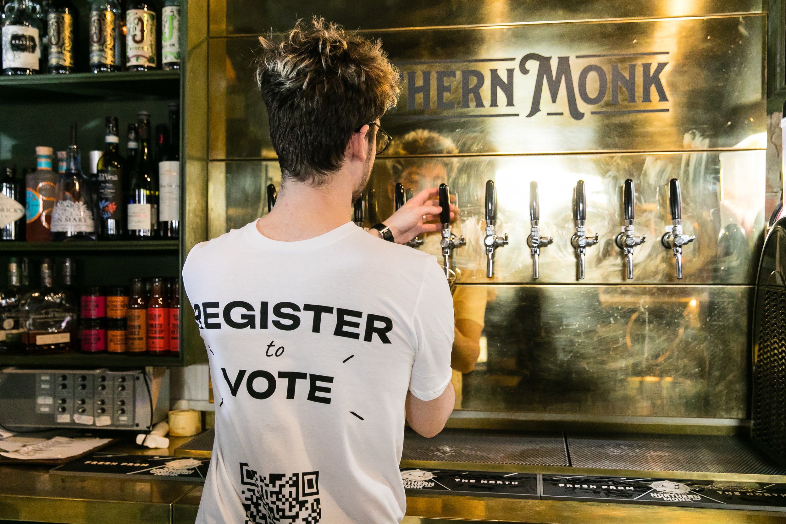 A man with glasses and a white T-shirt that says 'Register to Vote' is pouring a drink from a tap at a bar or brewery with six taps. The back of his T-shirt has a QR code. There are shelves with bottles and cans of drinks behind him.