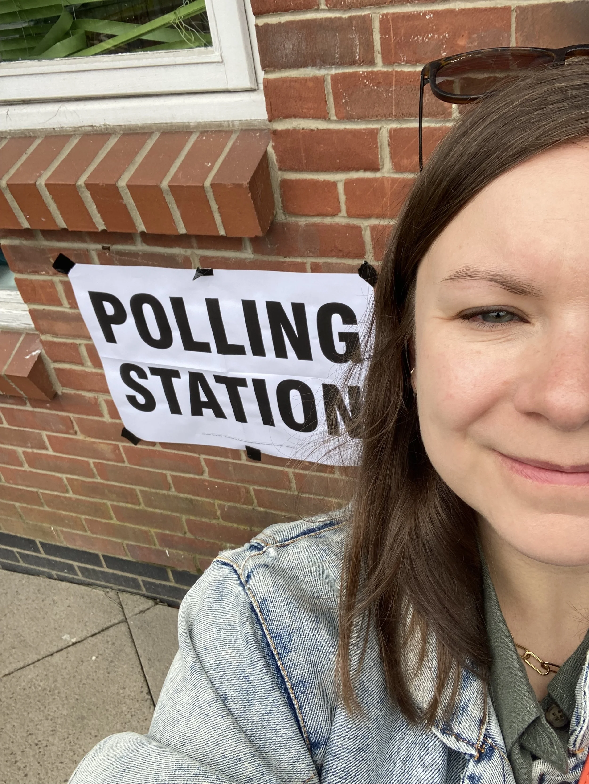 A woman taking a selfie in front of a polling station sign attached to a brick wall, with part of her face visible on the right side of the image.