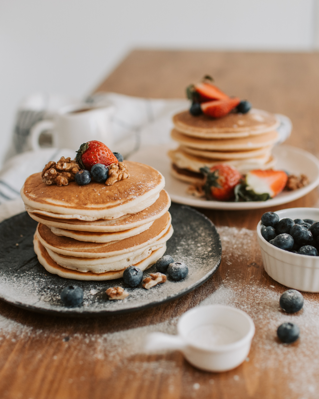 Stack of five pancakes topped with strawberries, blueberries, and walnuts, with additional strawberries and blueberries on a white plate and nearby. Pouring cream or syrup in a small white pitcher in the foreground.