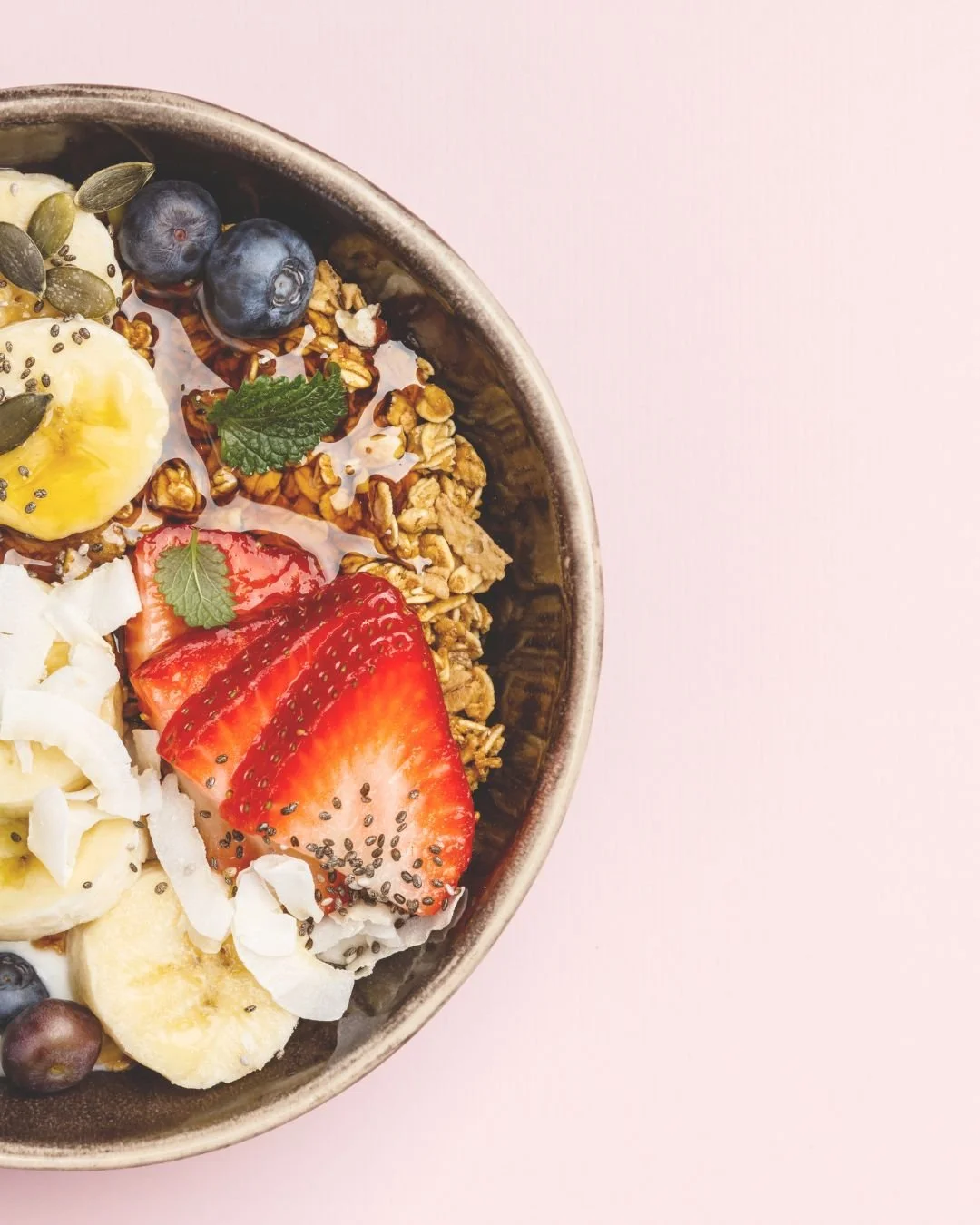 Bowl of granola topped with sliced strawberries, bananas, blueberries, shredded coconut, coconut flakes, pumpkin seeds, chia seeds, and fresh mint on a pink background. Sweet nourishing plant based breakfast.
