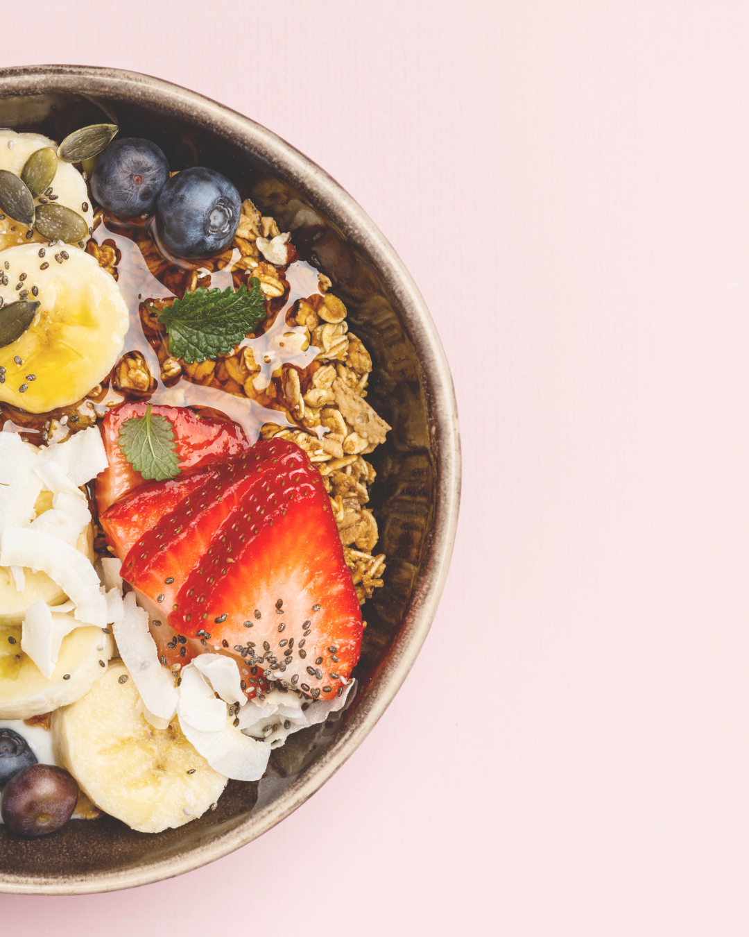Bowl of granola topped with sliced strawberries, bananas, blueberries, shredded coconut, coconut flakes, pumpkin seeds, chia seeds, and fresh mint on a pink background.