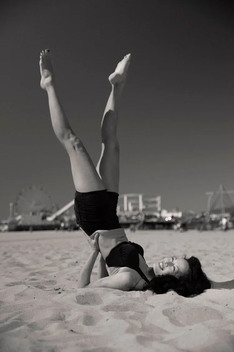 A woman performing an acrobatic yoga pose on the beach with her legs extended upward, wearing a black top and shorts, smiling with her eyes closed, in black and white. In the background, there is a Ferris wheel and other amusement park rides.