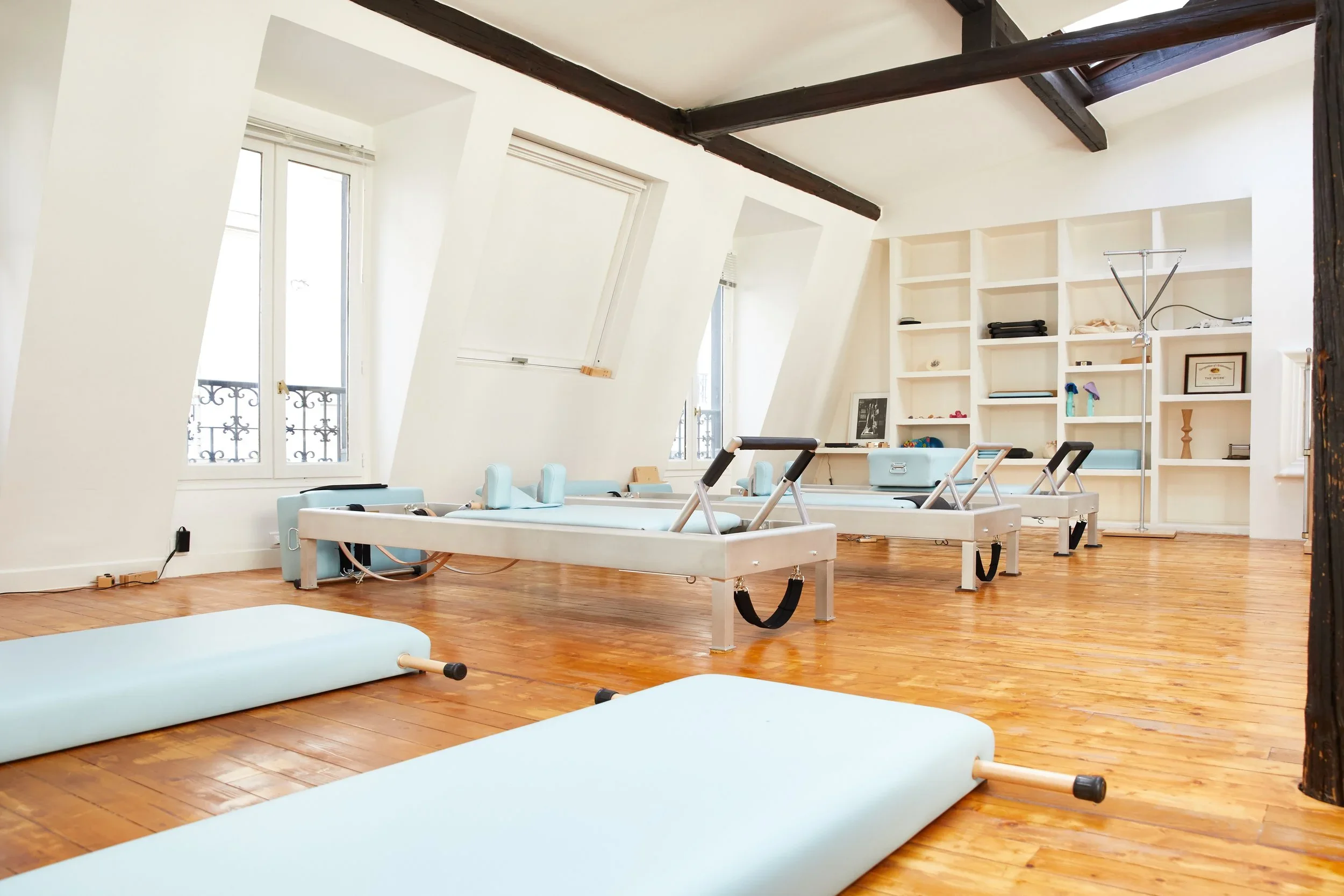 A spacious Pilates studio with white walls, wooden floors, large windows, and Pilates reformer machines, mats, and exercise equipment.