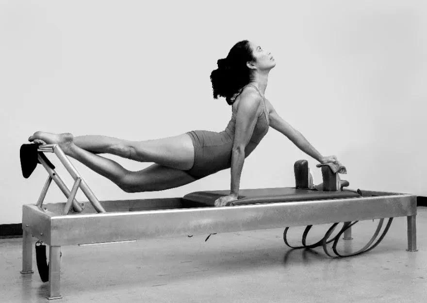 A woman practicing yoga on a specialized Pilates reformer machine in a studio with a plain background.