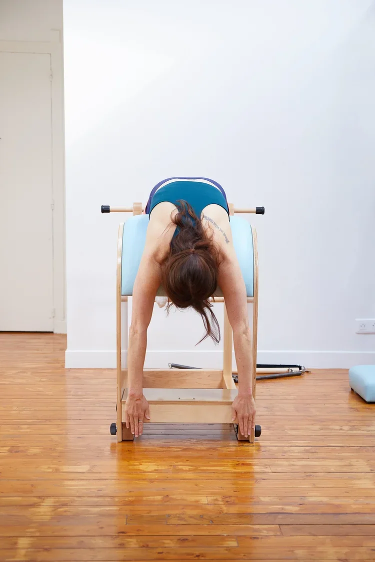 Woman exercising on a Pilates reformer machine in a studio with wooden floors and white walls.