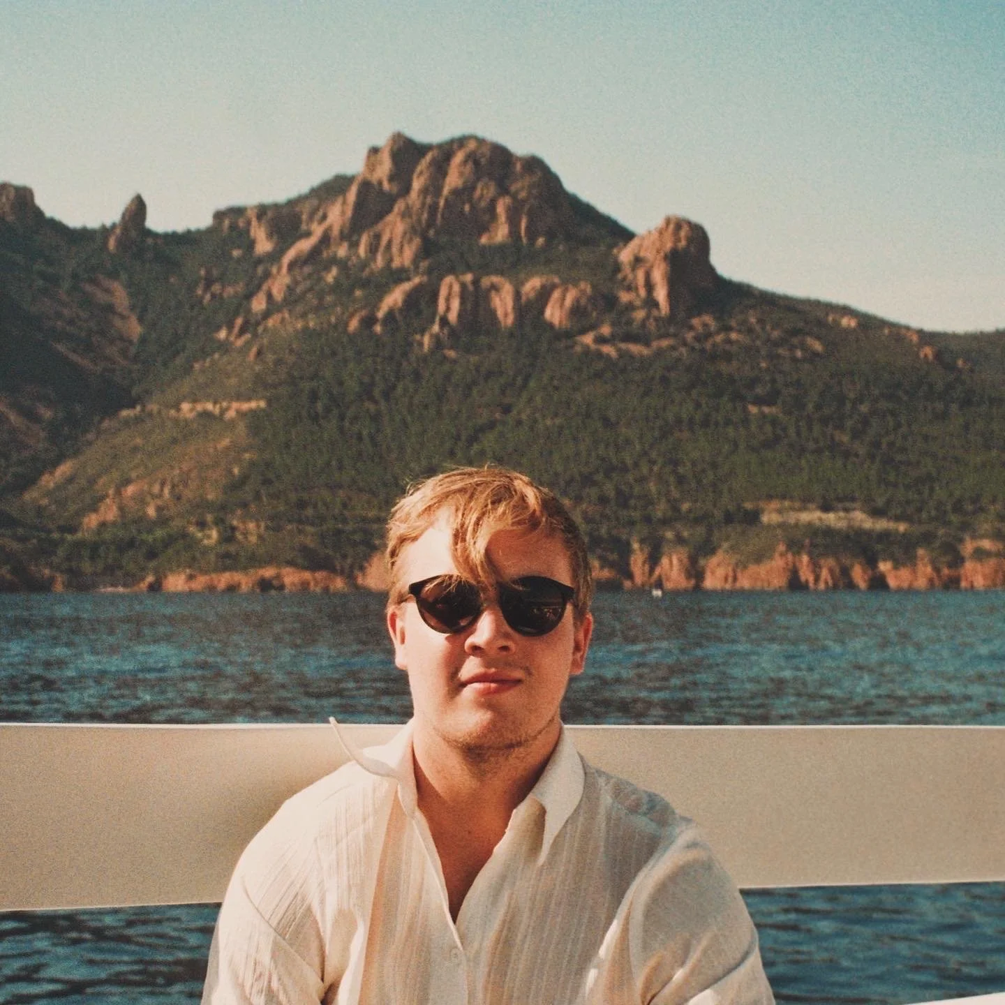 A young man wearing sunglasses and a light-colored button-up shirt, sitting on a boat to St Tropez with a mountain and lake in the background during daytime.