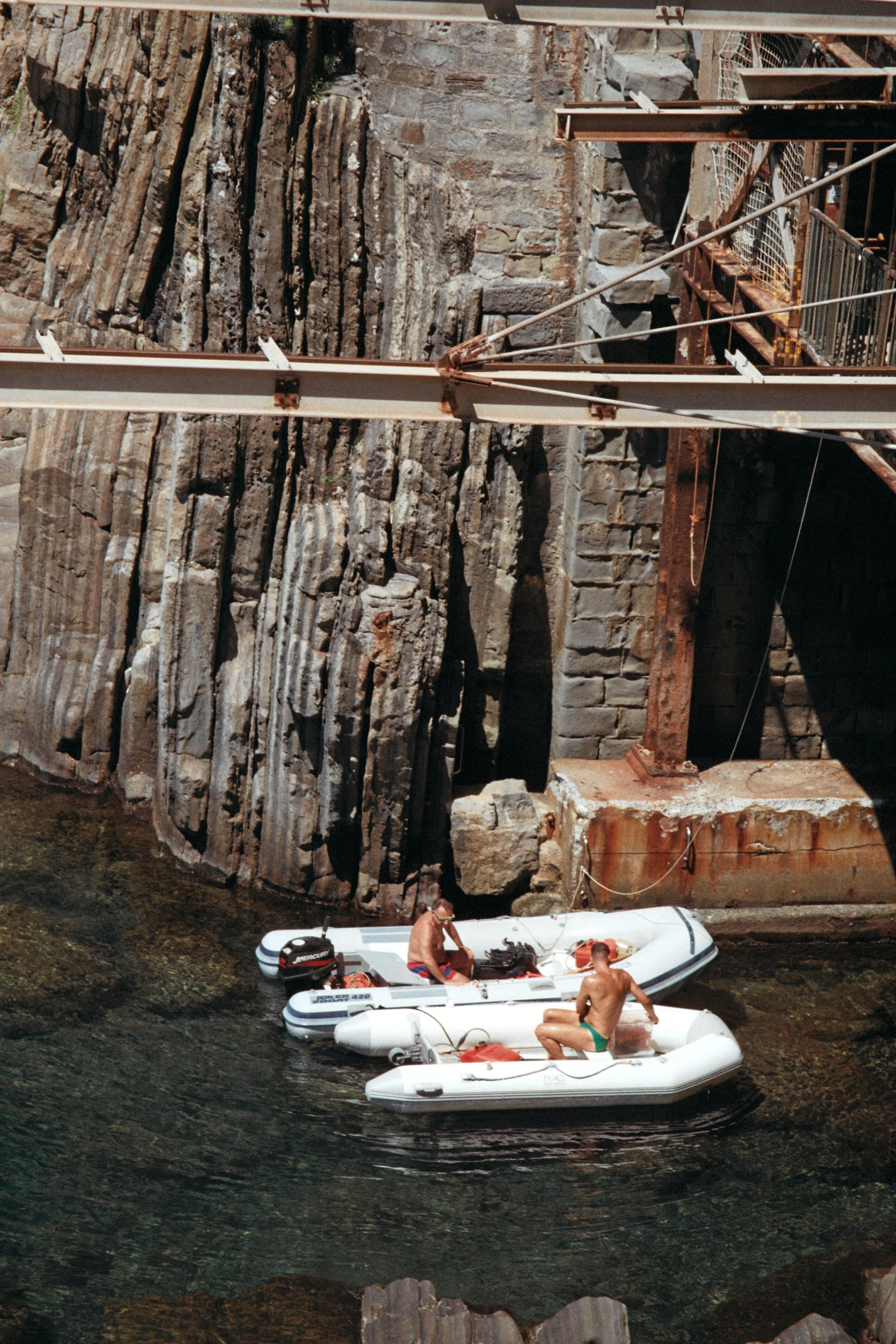 Two men in swimsuits on inflatable boats near a rocky shoreline under a bridge with rusted steel beams and stone pillar supports.