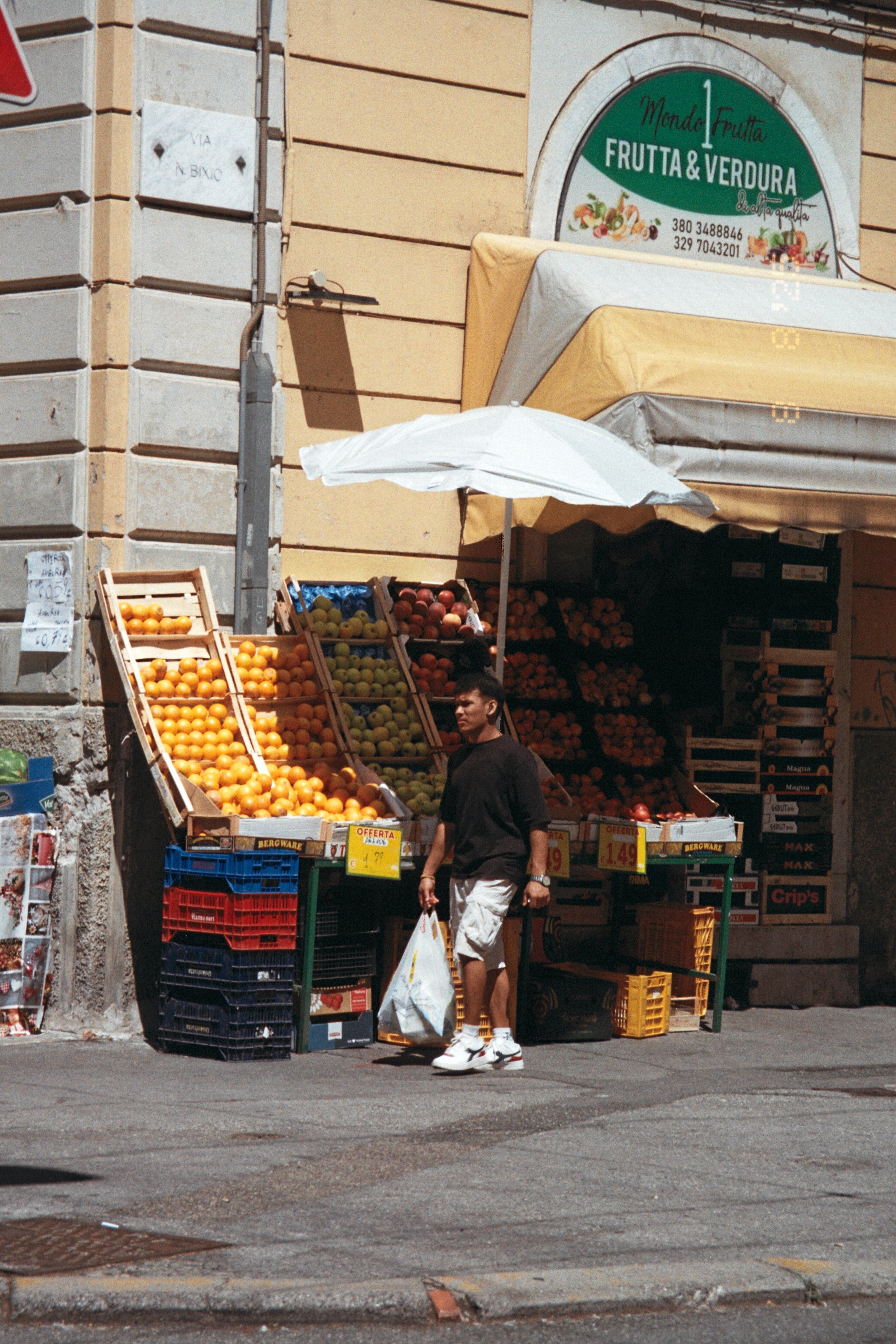 A man walking past a street produce stand with oranges and apples, with a sign reading 'Frutta & Verdura' above.